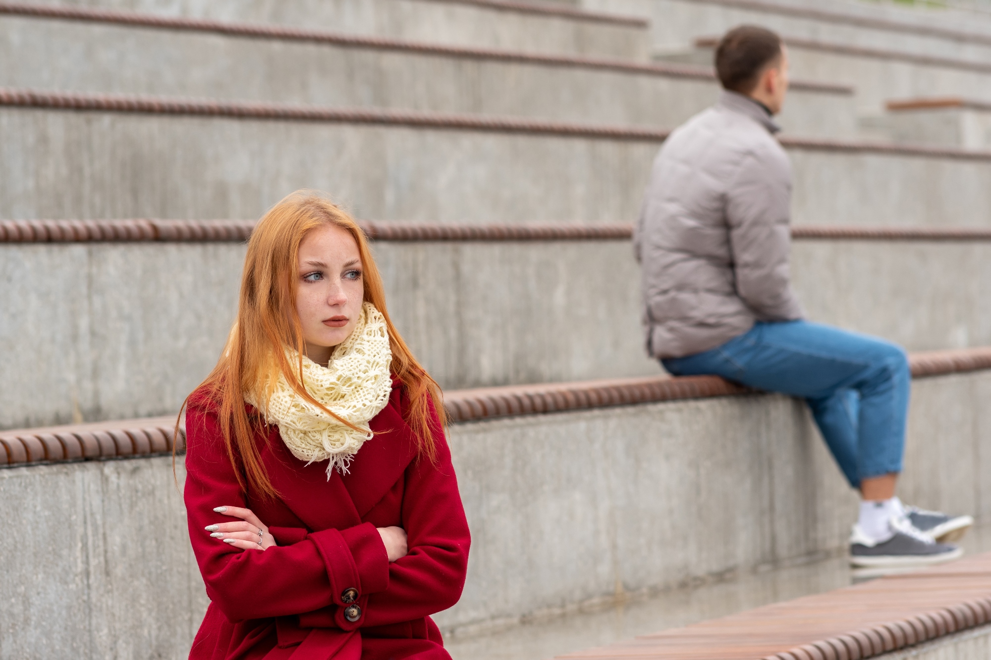 A woman with red hair wearing a red coat and a cream scarf sits on concrete steps with her arms crossed, looking away thoughtfully. In the background, a man in a gray jacket and blue jeans sits on the steps, facing away from her.