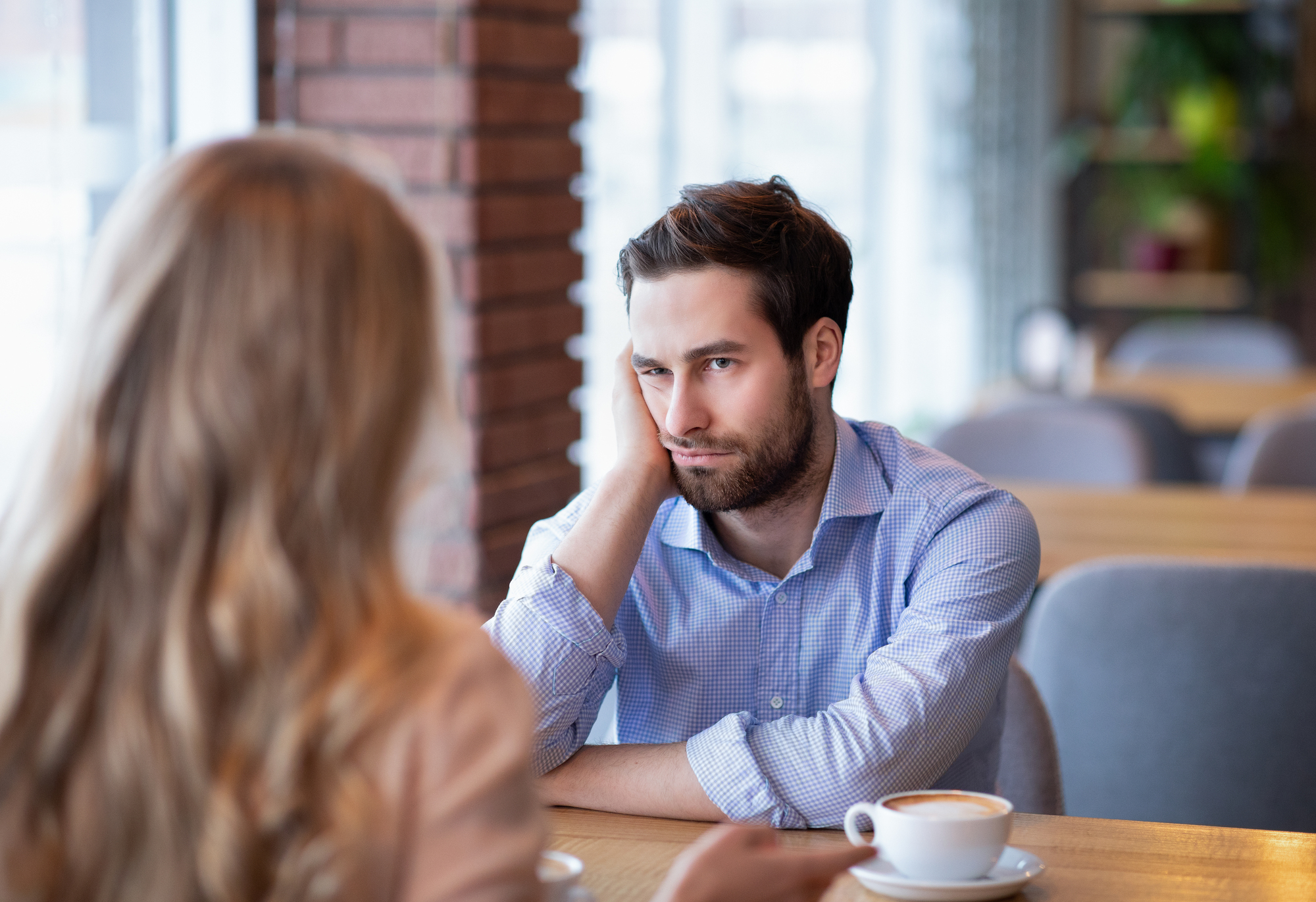 A man with a beard, wearing a blue shirt, sits at a table in a café, resting his head on his hand. He has a serious expression and is looking at a woman with long blonde hair sitting across from him. Both have coffee cups in front of them.