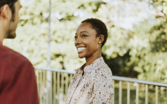 A person with short hair smiles at another person outdoors during daytime. They are wearing a patterned shirt and standing near a railing with trees in the background.