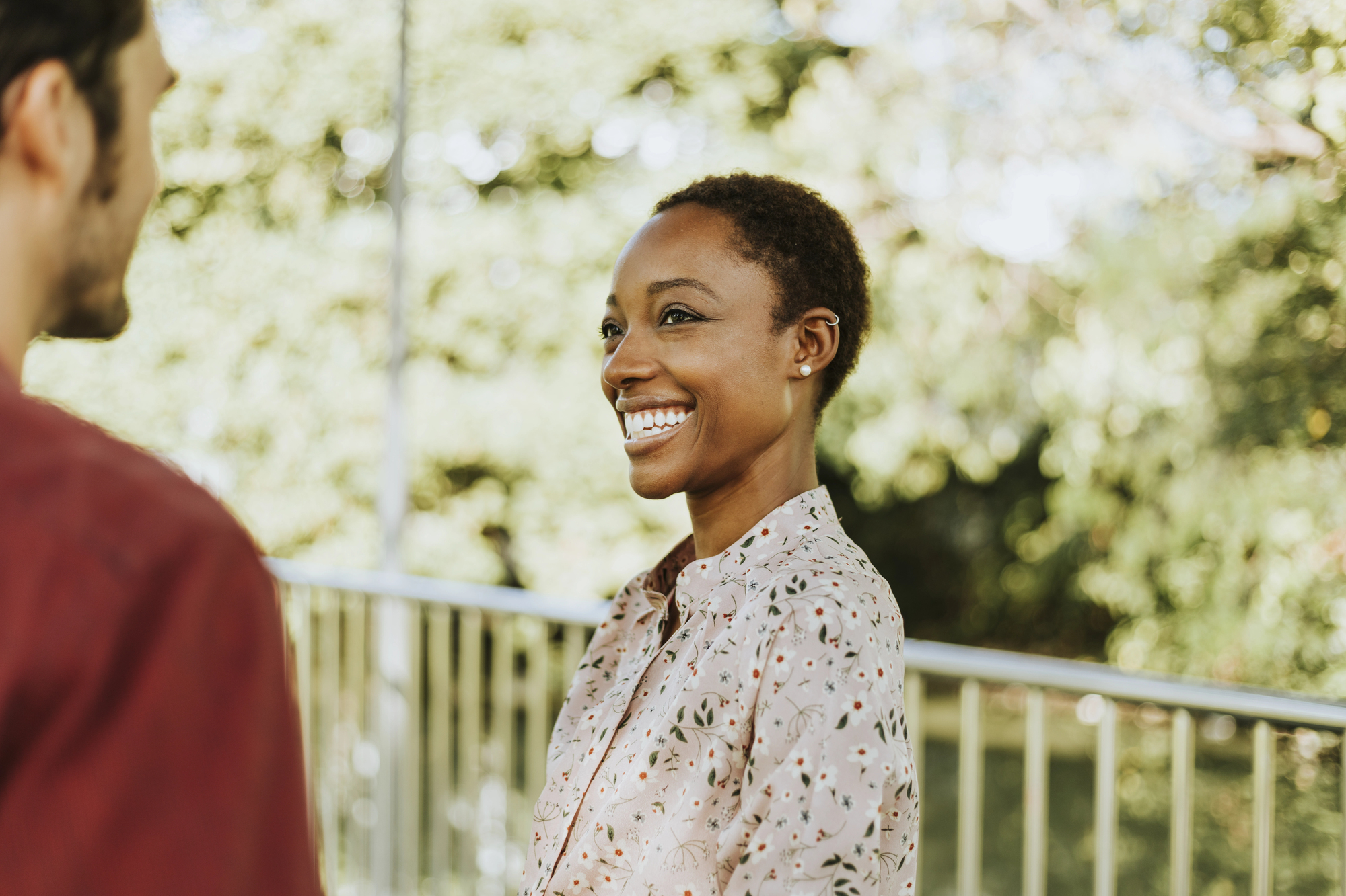 A person with short hair smiles at another person outdoors during daytime. They are wearing a patterned shirt and standing near a railing with trees in the background.