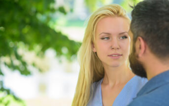 A woman with long blonde hair and a nose piercing stands outdoors, facing a man with a beard. Bright green leaves are visible in the background. Both individuals appear to be engaged in a conversation.