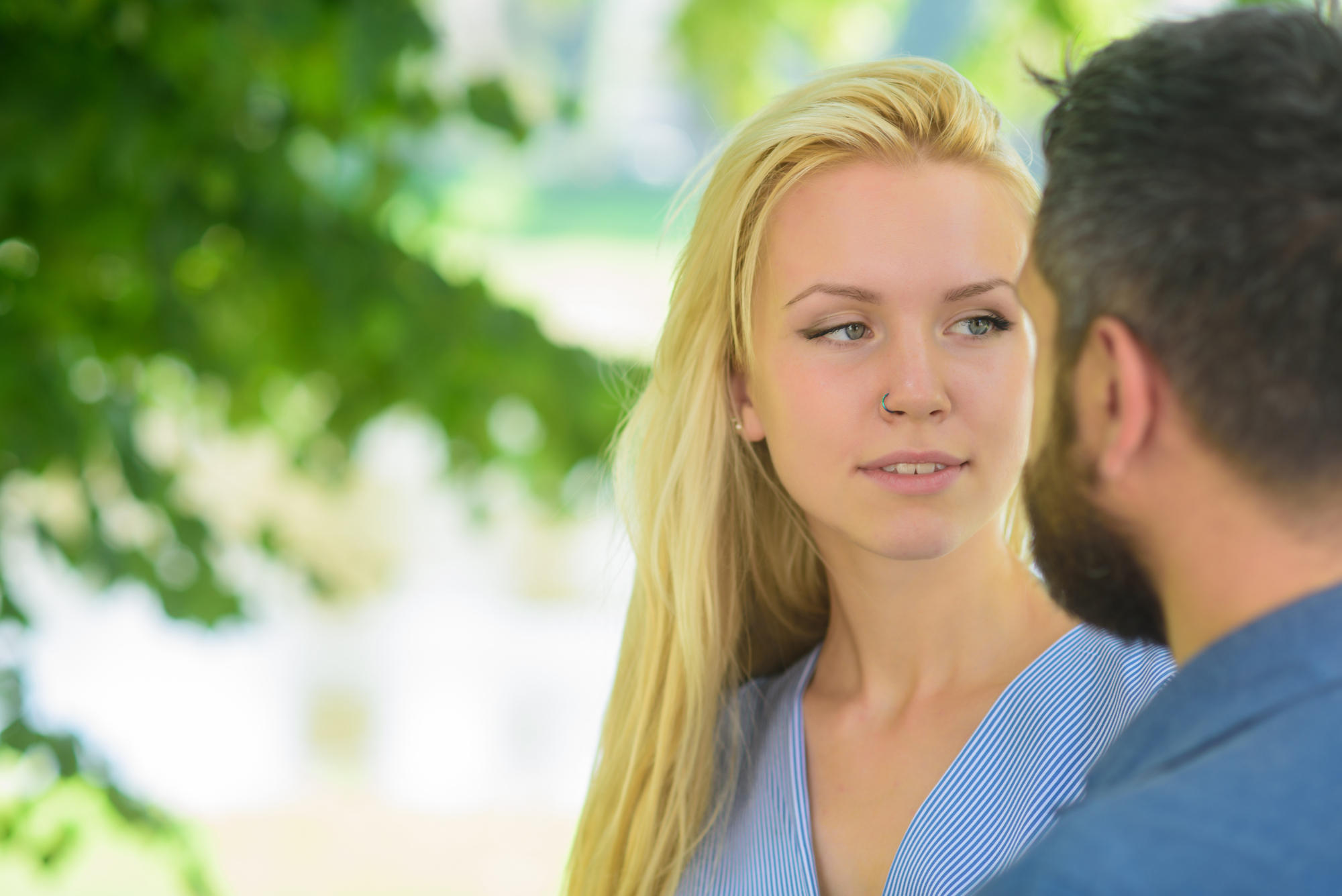 A woman with long blonde hair and a nose piercing stands outdoors, facing a man with a beard. Bright green leaves are visible in the background. Both individuals appear to be engaged in a conversation.