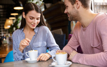 A woman and a man are sitting at a table in a cozy café, each with a cup of coffee. The woman, dressed in a blue striped shirt, is stirring her coffee while smiling. The man, wearing a pink shirt, is looking at her with a smile.