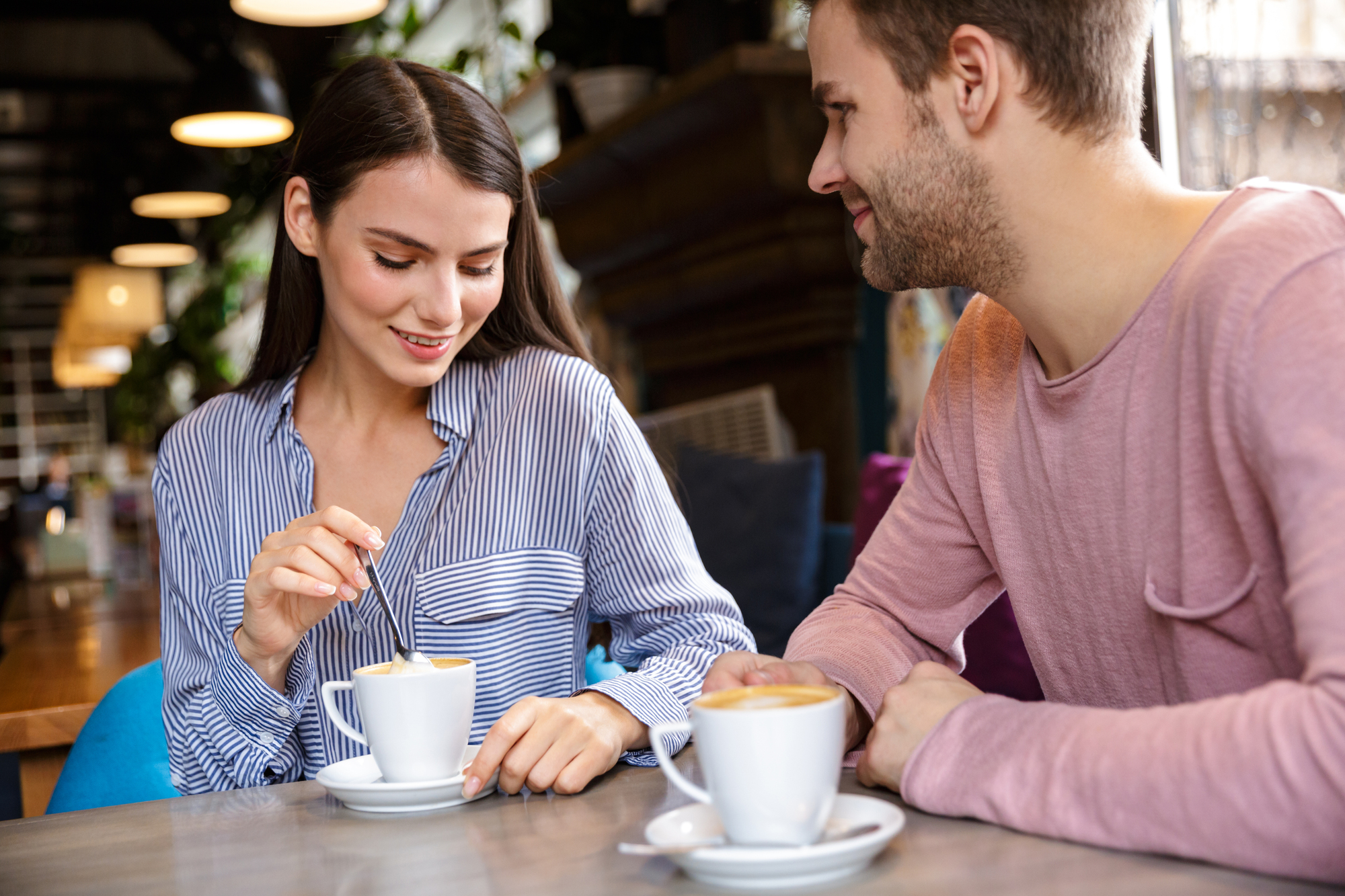 A woman and a man are sitting at a table in a cozy café, each with a cup of coffee. The woman, dressed in a blue striped shirt, is stirring her coffee while smiling. The man, wearing a pink shirt, is looking at her with a smile.