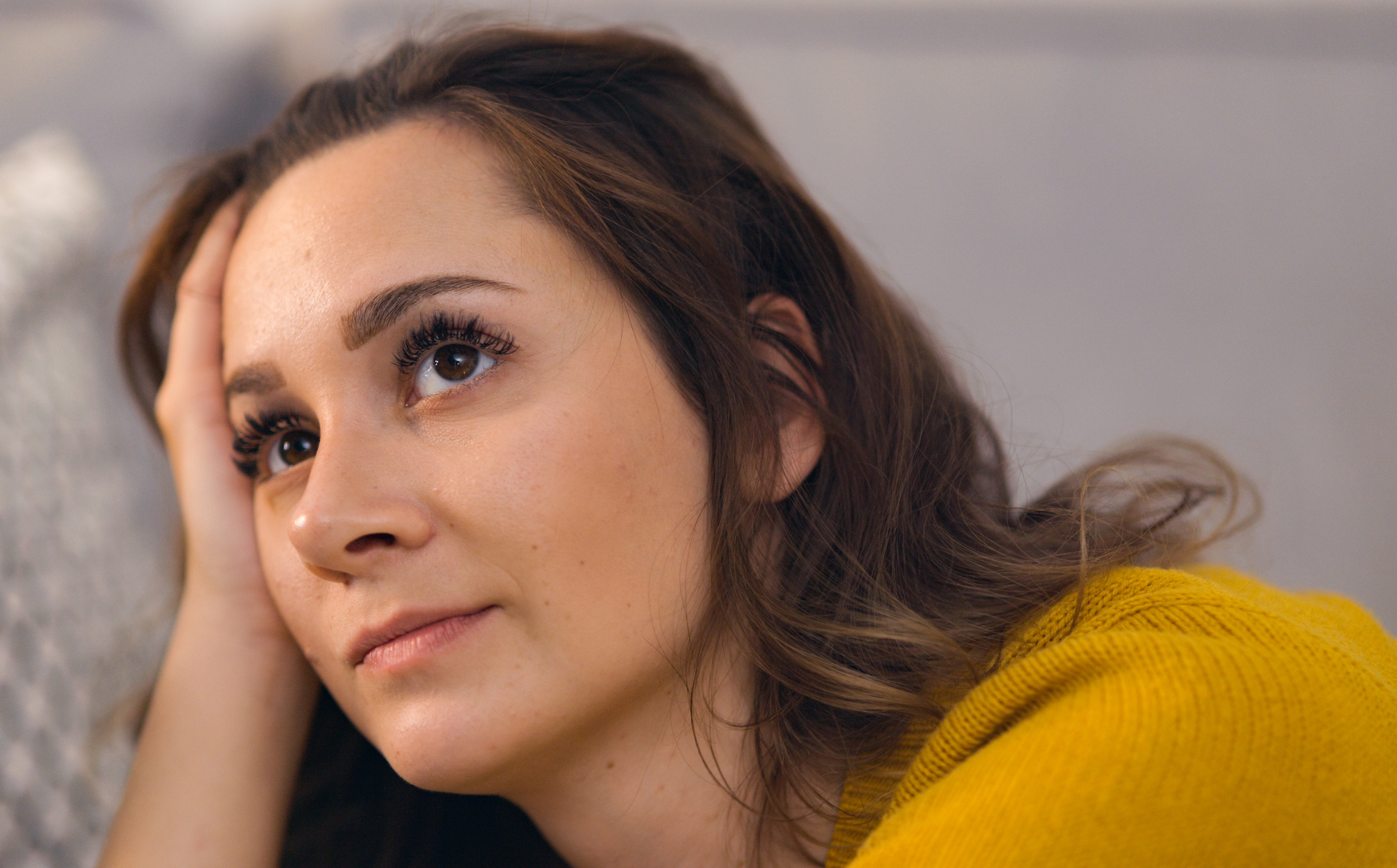 A thoughtful woman with long eyelashes and brown hair rests her head on her hand, looking slightly upwards. She is wearing a mustard yellow sweater and appears deep in thought with a calm and relaxed expression.