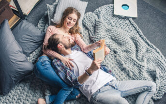 A couple is lying on a gray knitted rug, smiling and holding a book. The woman, in a pink sweater, has her arm around the man, who is wearing a checkered shirt over a white t-shirt and jeans. They are surrounded by pillows and appear to be relaxed and happy.