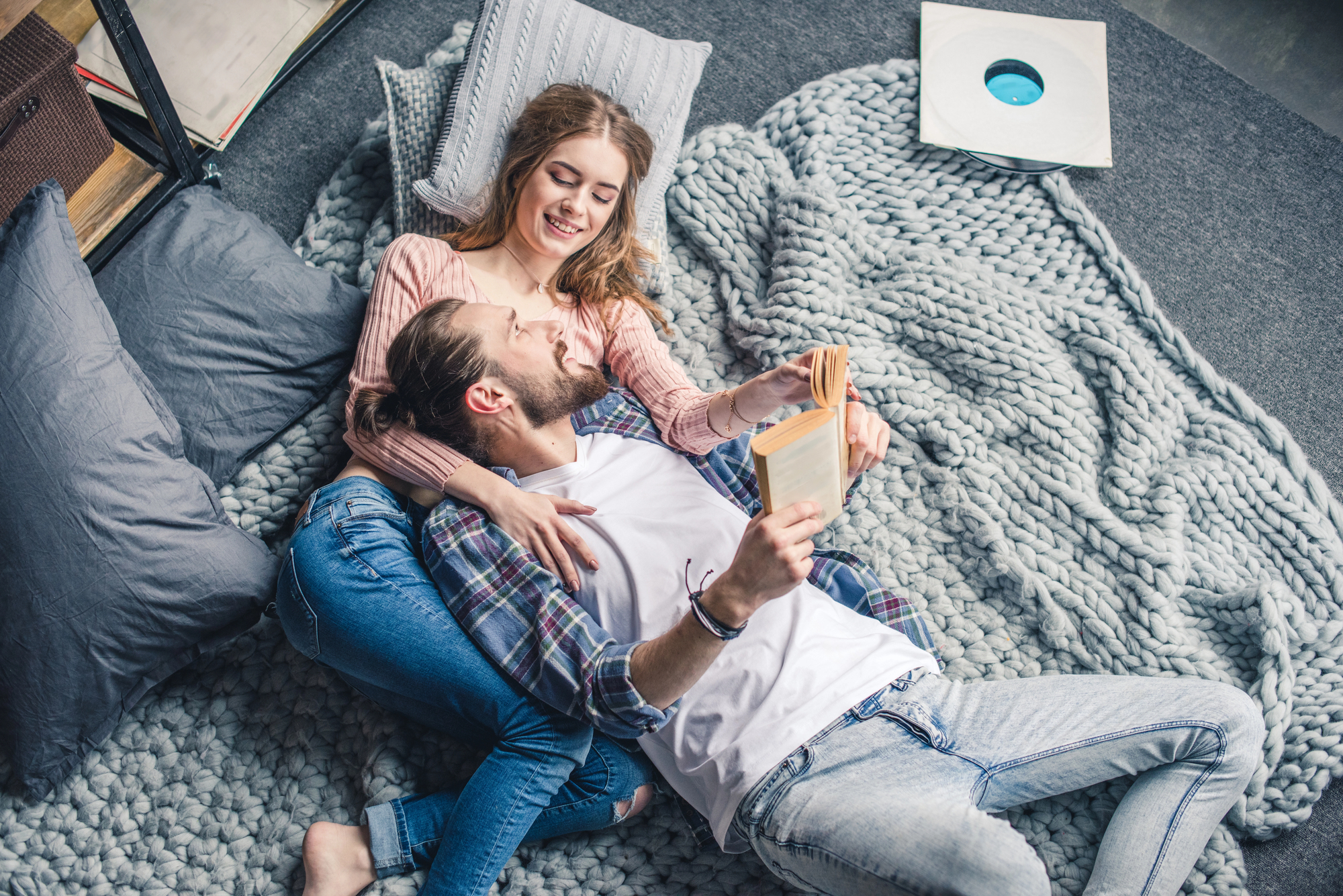 A couple is lying on a gray knitted rug, smiling and holding a book. The woman, in a pink sweater, has her arm around the man, who is wearing a checkered shirt over a white t-shirt and jeans. They are surrounded by pillows and appear to be relaxed and happy.