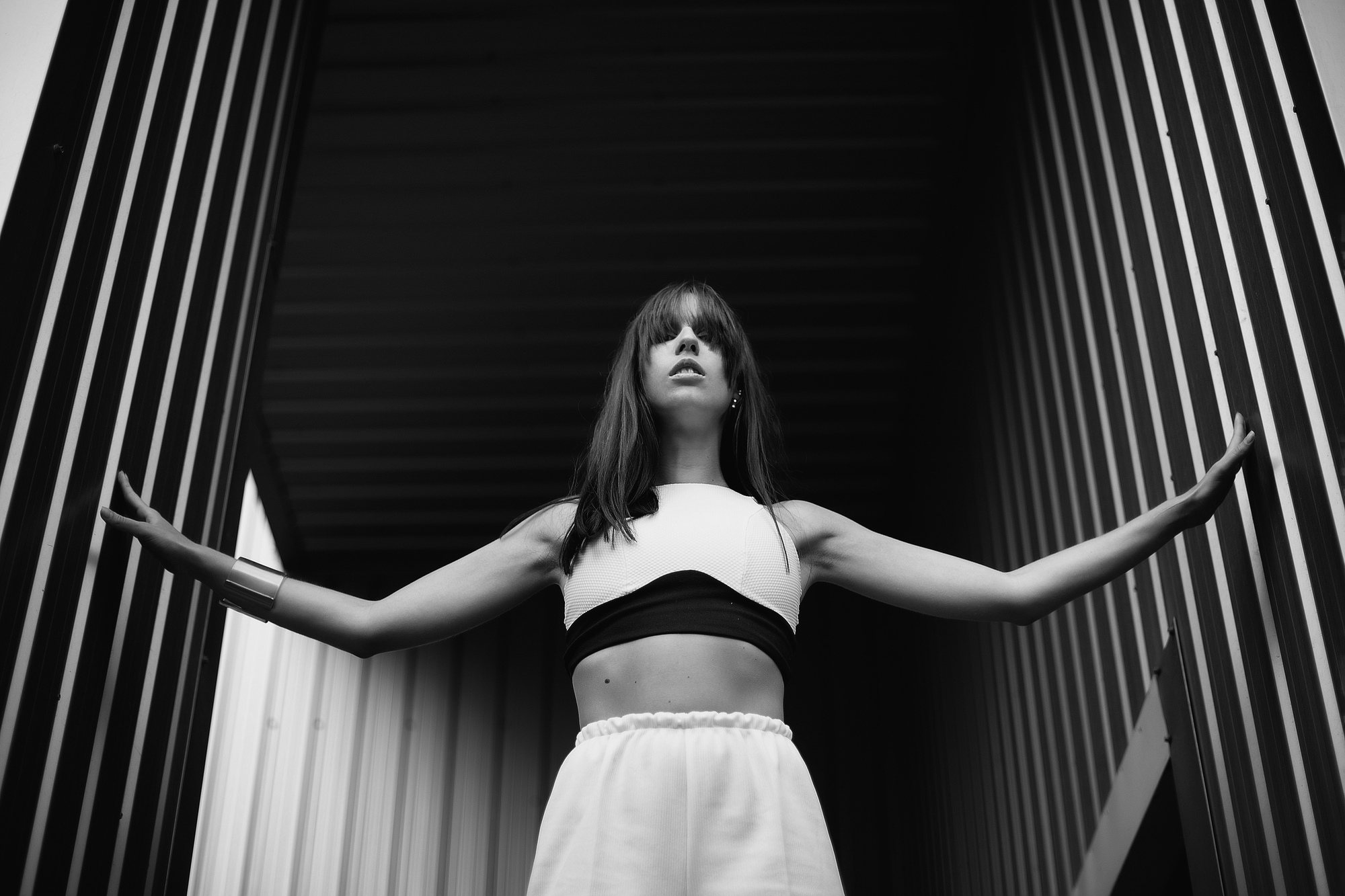 A woman stands confidently with arms outstretched, touching the sides of a narrow, vertical structure. She wears a sporty outfit with a white crop top and high-waisted pants. The image is in black and white, highlighting the contrast and the dramatic, moody atmosphere.