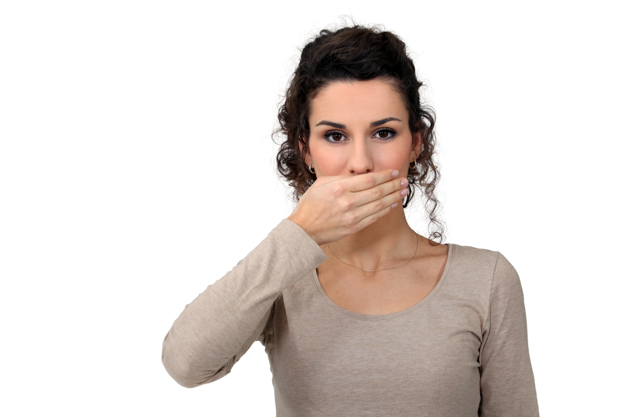 A woman with curly hair wearing a light brown long-sleeve shirt is holding her hand over her mouth. She is looking directly at the camera against a plain white background.