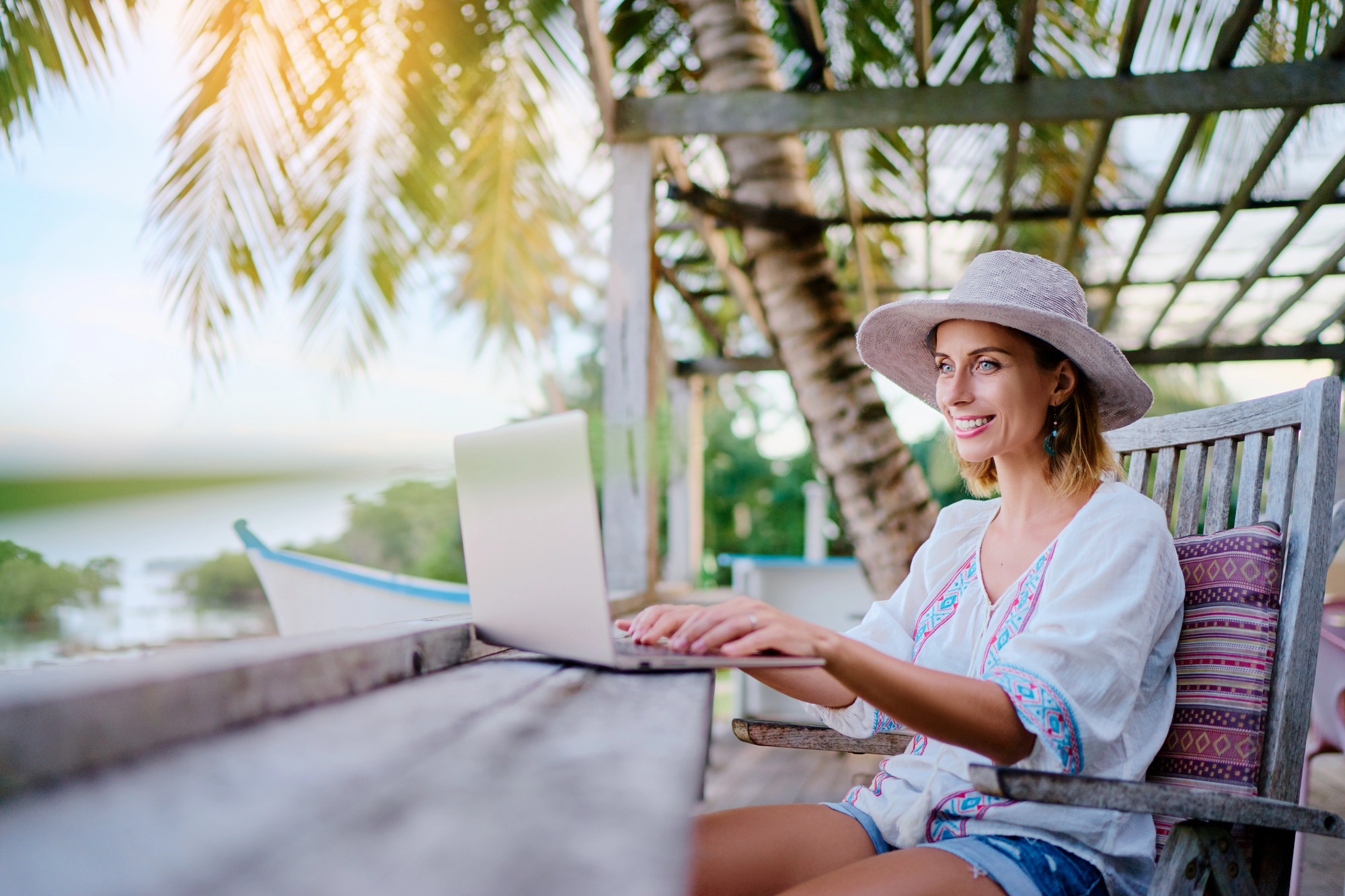 A woman in a white blouse and sunhat sits at an outdoor table under a palm tree, typing on a laptop. She smiles while working, with a serene tropical background of a beach and lush greenery.