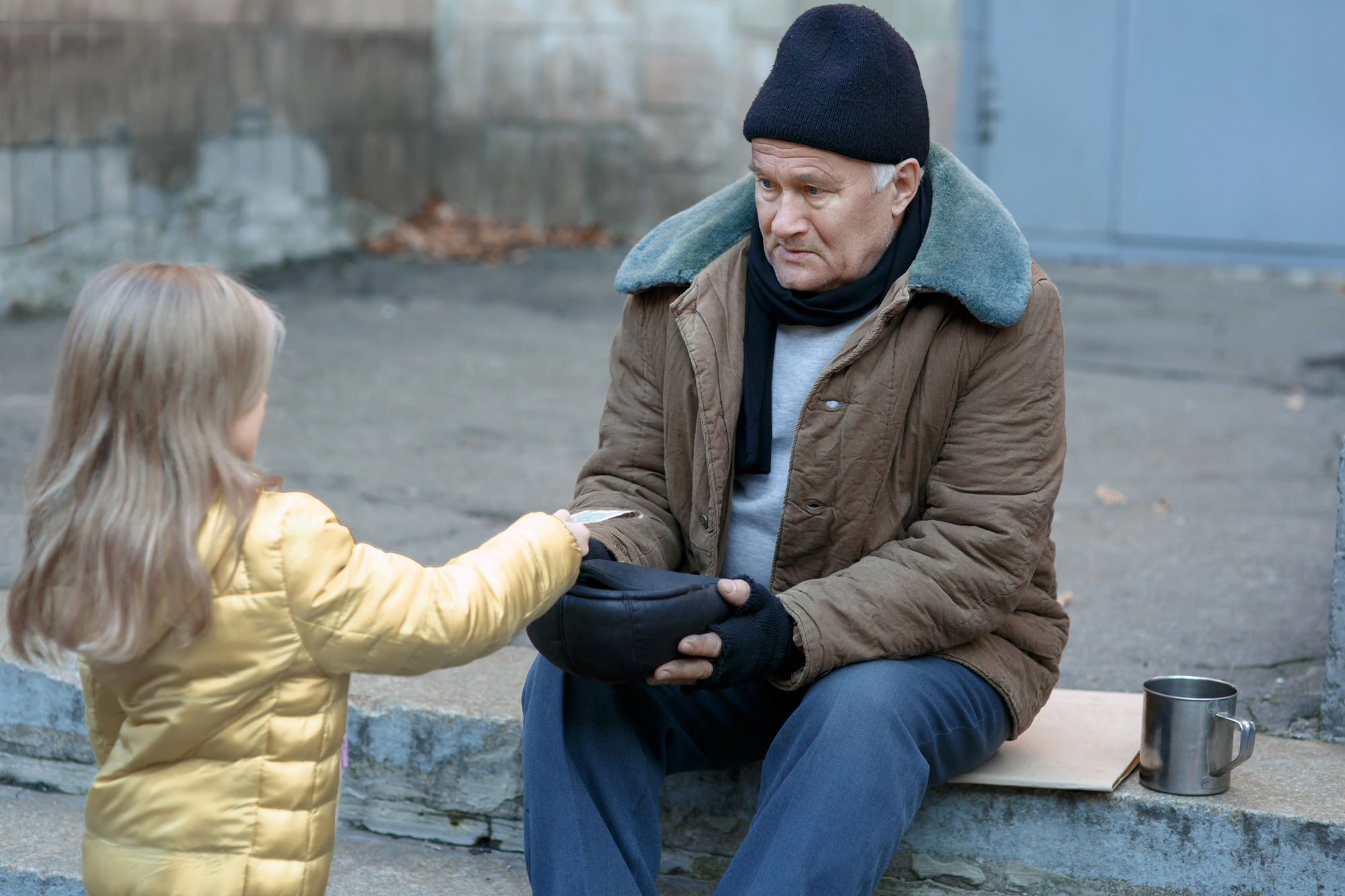 A young girl in a yellow jacket offers a banknote to an elderly man sitting on a step, holding a hat. The man, dressed in a brown coat and black beanie, looks at the girl with a warm expression. A metal cup sits beside him on the ground.