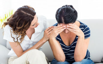 A woman with dark hair wearing a striped shirt sits on a coach, holding her head in her hands in distress. Another woman with light hair in a white blouse sits next to her, offering comfort by placing a hand on her shoulder and looking at her empathetically.