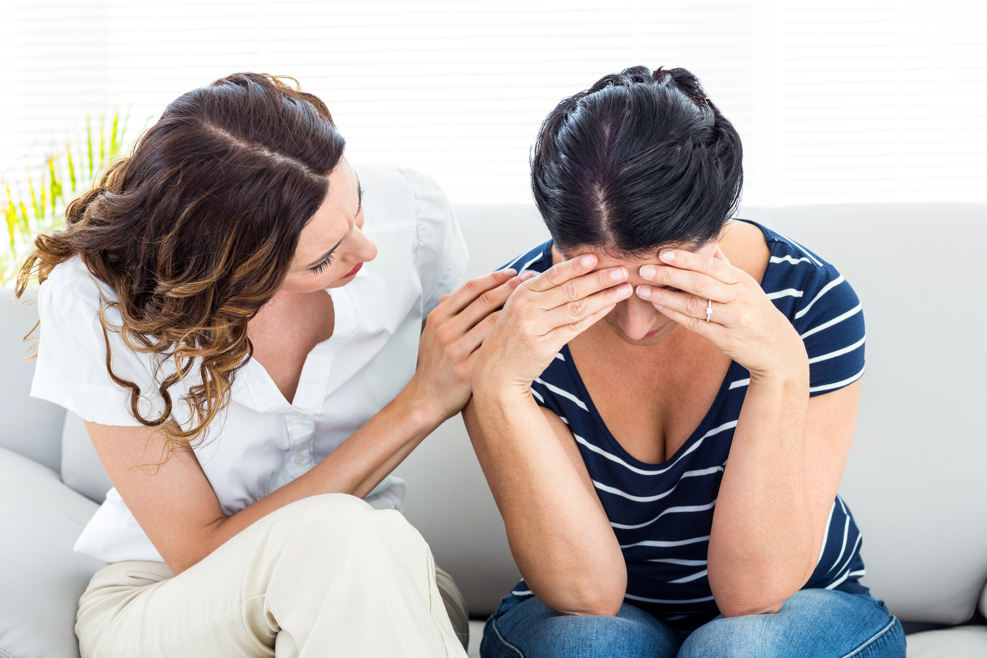 A woman with dark hair wearing a striped shirt sits on a coach, holding her head in her hands in distress. Another woman with light hair in a white blouse sits next to her, offering comfort by placing a hand on her shoulder and looking at her empathetically.