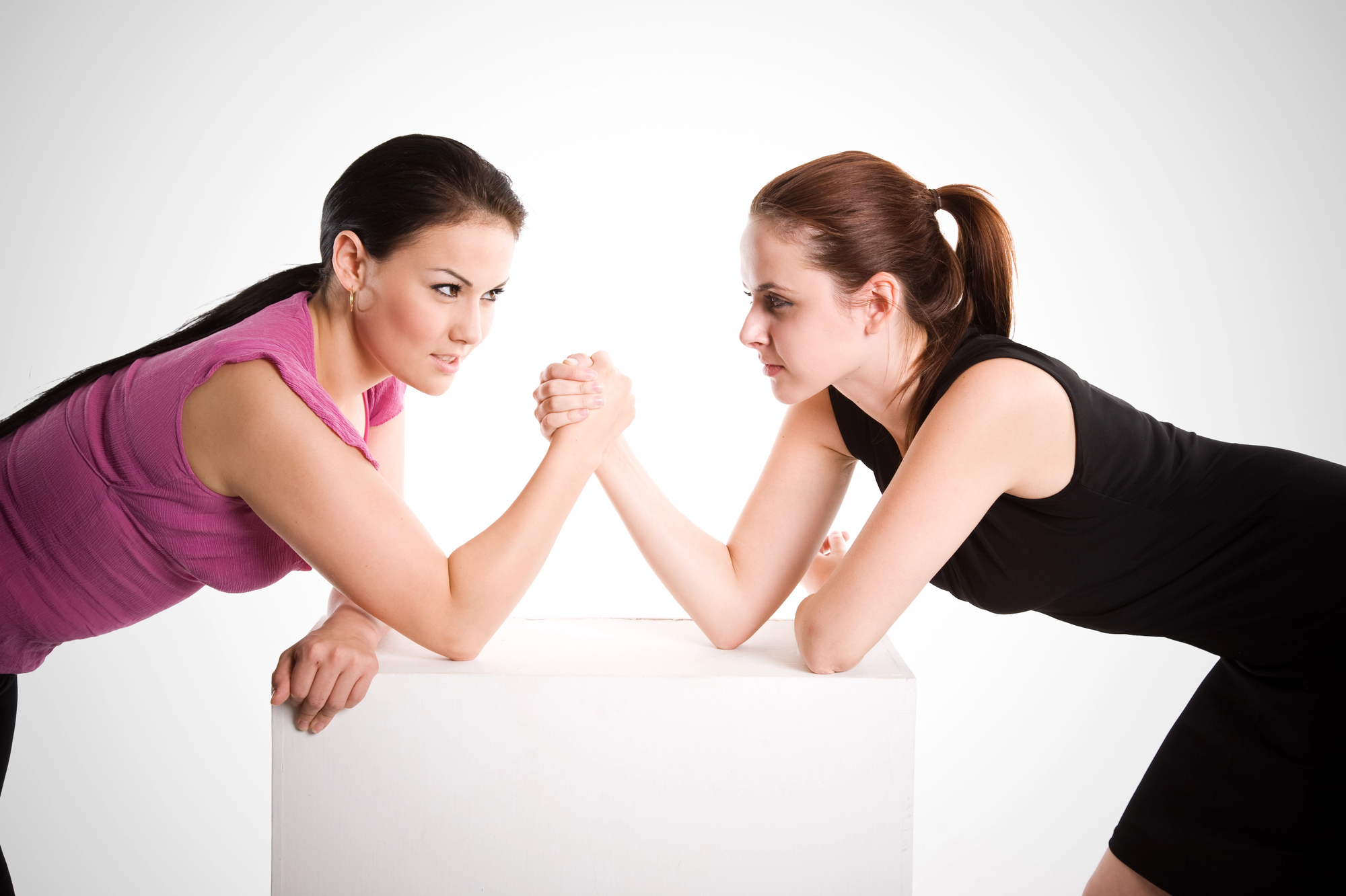 Two women are engaged in an arm wrestling match, each leaning over a white rectangular block. One woman is wearing a pink top, the other in a black top, and they both have focused expressions, intensely staring at each other as they grip hands.