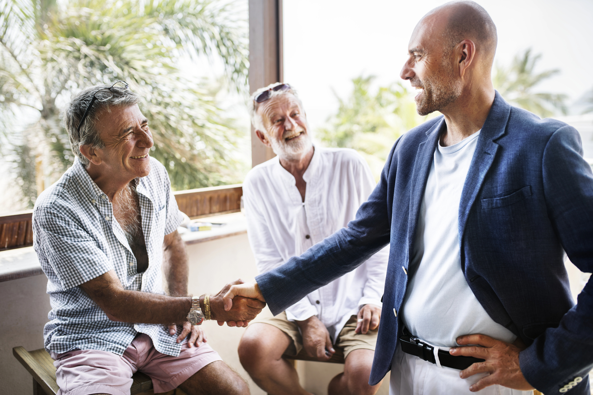 Three men socialize on a sunny porch with greenery in the background. One man in a blue blazer and light shirt shakes hands with a man in a checkered shirt and pink shorts, while another man in white shirt and shorts, wearing sunglasses, looks on.