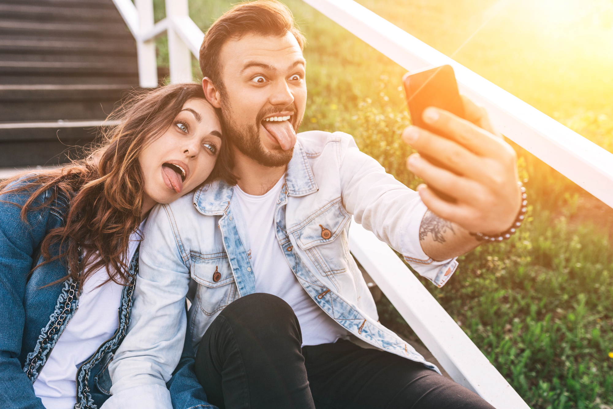 A young man and woman sit on outdoor steps, posing for a selfie with playful facial expressions. The man holds the phone, sticking his tongue out, while the woman leans on his shoulder, making a funny face. They both wear casual denim jackets.