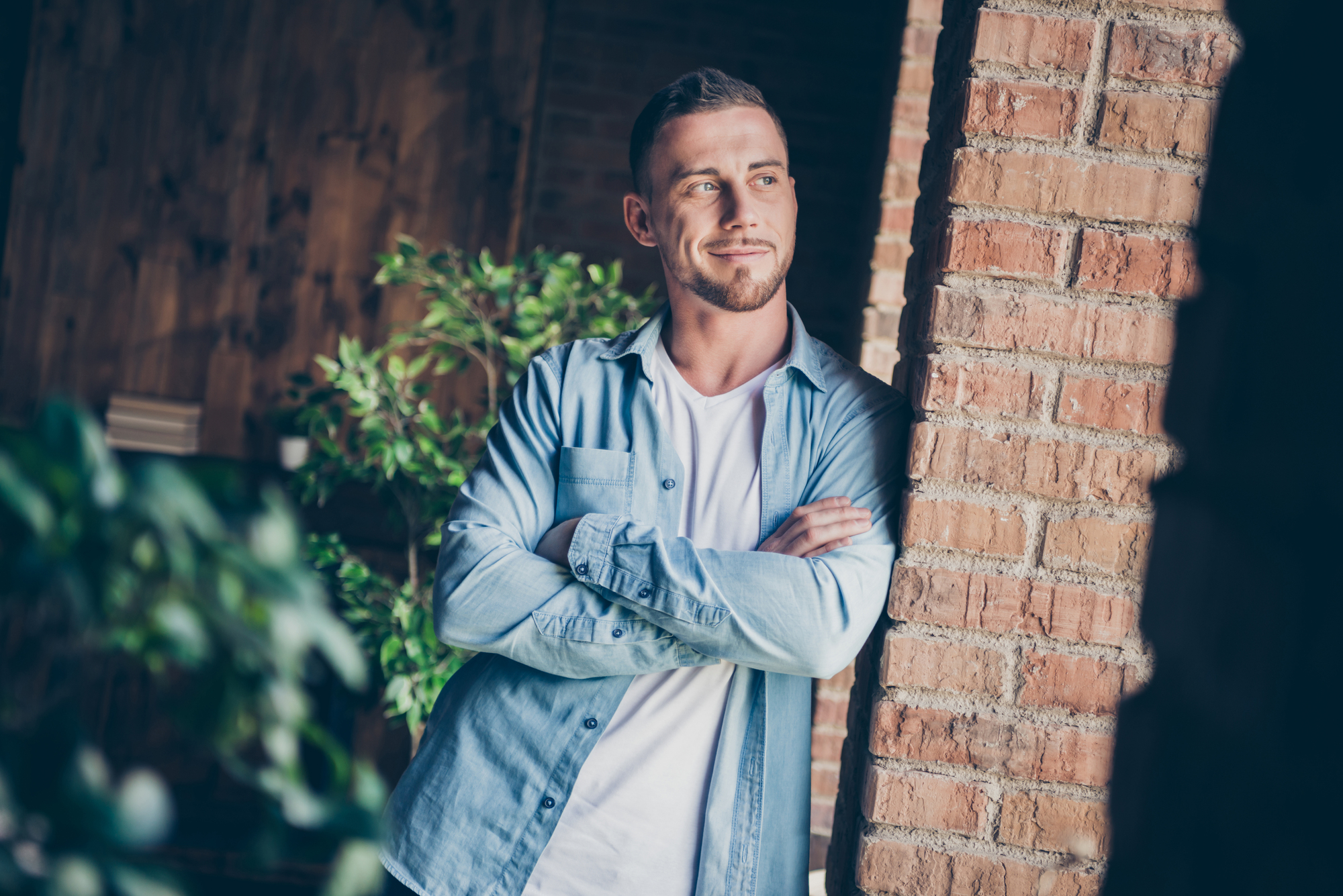 A man with short hair and a slight beard standing indoors with his arms crossed, leaning against an exposed brick wall. He is wearing a white t-shirt under a light blue denim shirt. In the background, there is a plant and wooden decor.