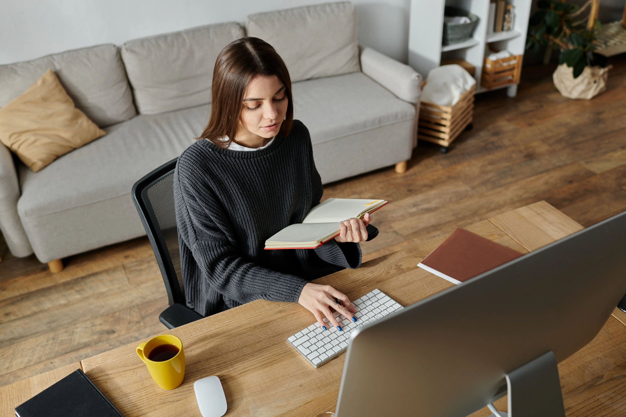 A person sits at a desk working on a computer while holding an open book. They are wearing a dark sweater and the desk also has a yellow mug, a closed notebook, and a computer mouse. The background shows a couch and bookshelves in a cozy room.