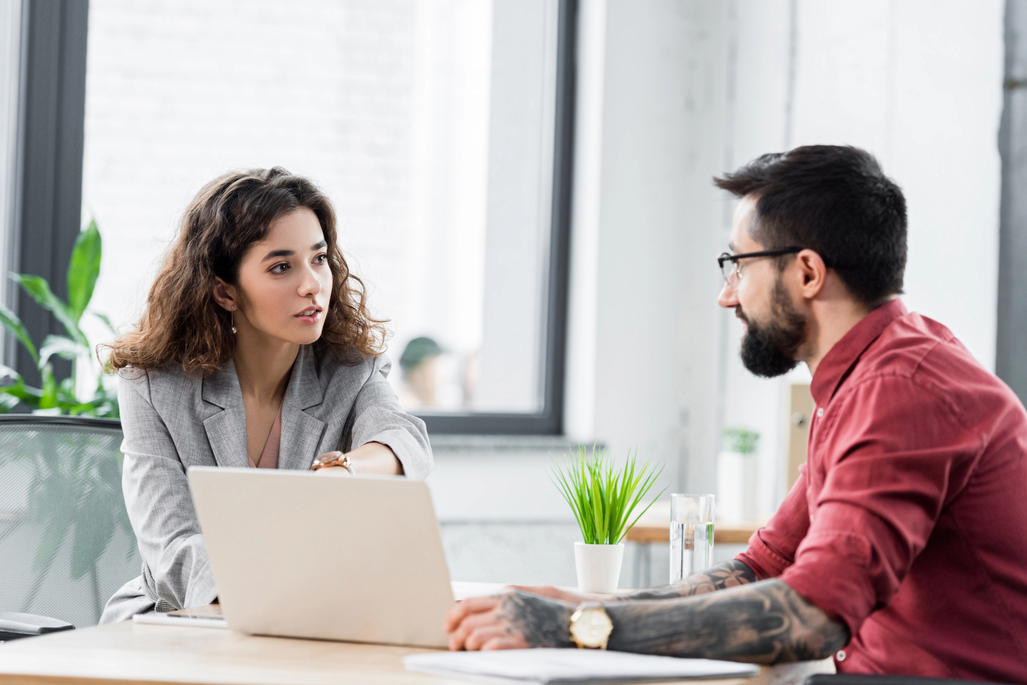 A woman in a gray blazer and a man with glasses, a beard, and arm tattoos are having a conversation at a desk in a modern office. They are facing each other with a laptop, small potted plant, and glass of water on the desk between them.