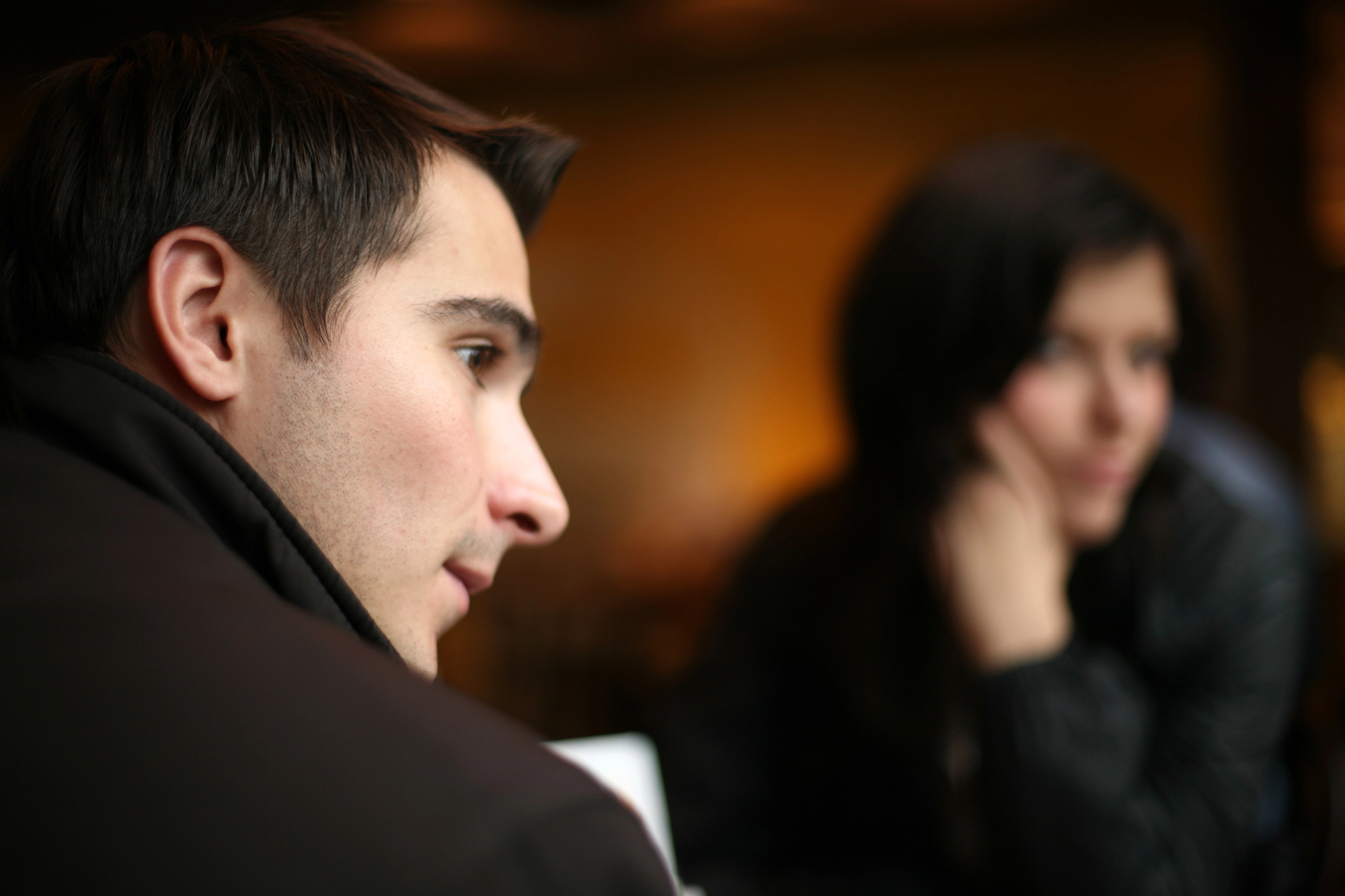 A young man in focus looks thoughtfully into the distance while a woman in the background, blurred, rests her chin on her hand. Both are indoors with a warm, softly lit ambiance.