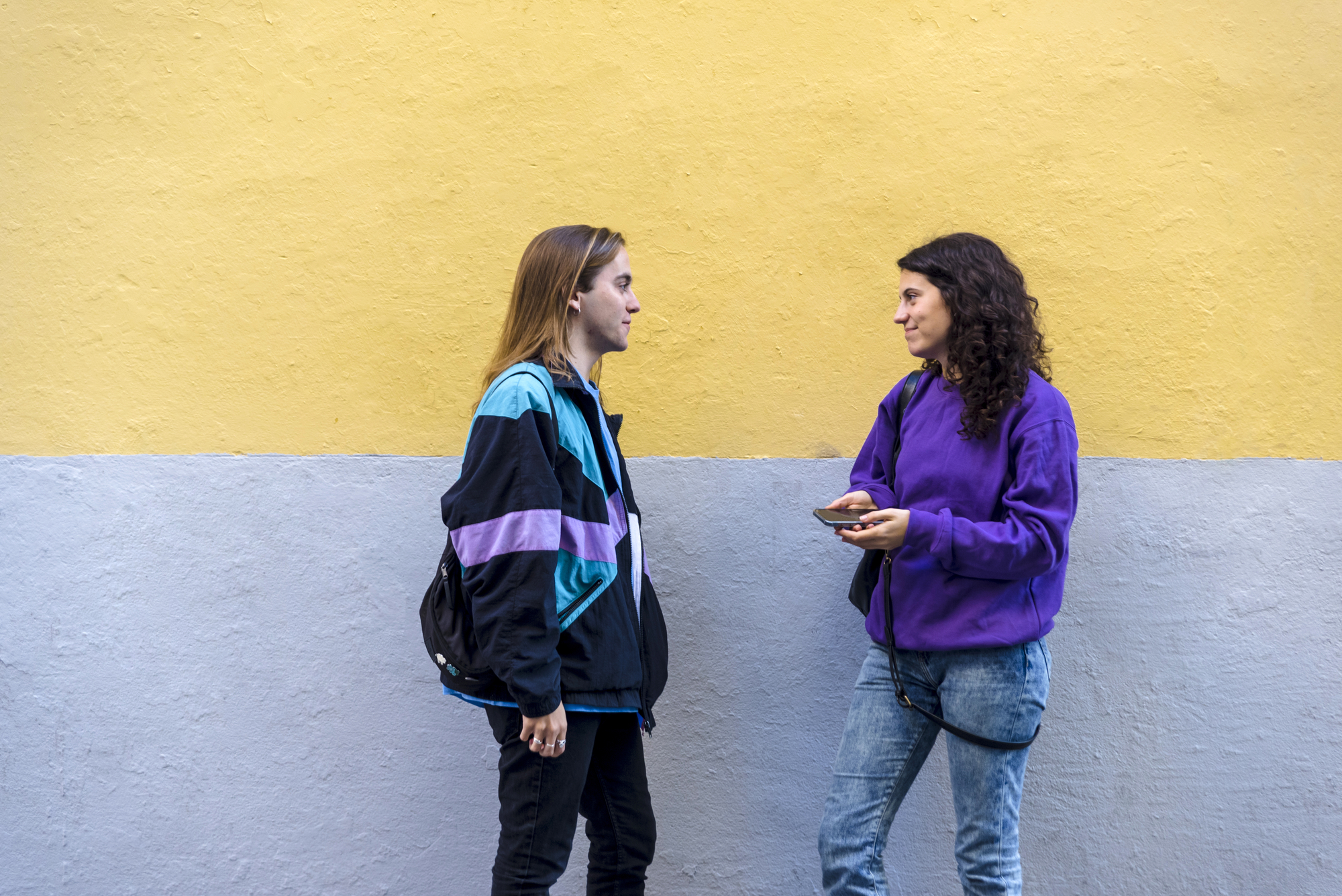 Two people are standing and engaging in conversation against a two-tone yellow and gray wall. One is wearing a blue and black jacket, and the other is dressed in a purple sweatshirt and jeans, holding a phone. Both appear relaxed and attentive.