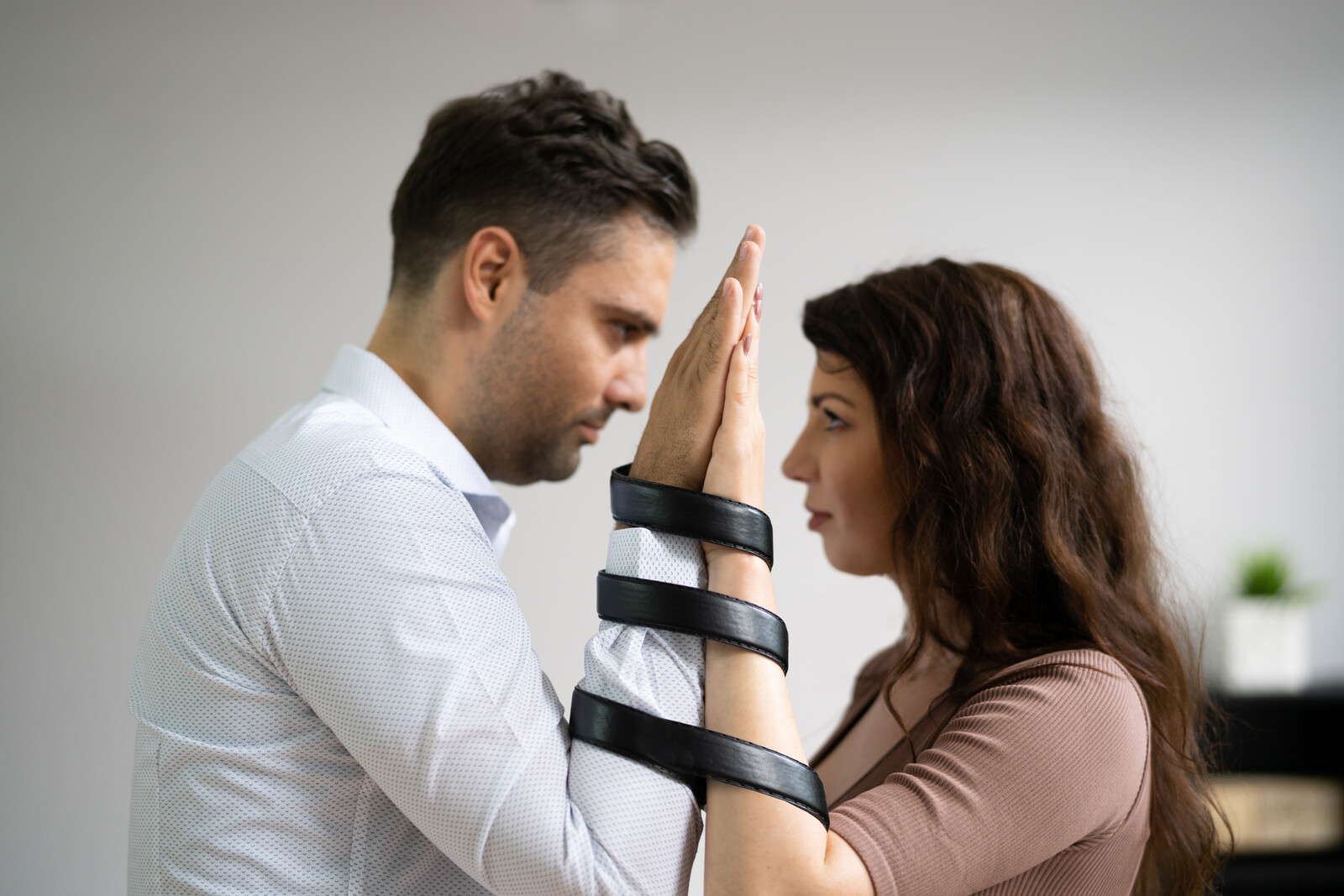 A man and a woman face each other, palms pressed together, with black straps binding their arms. The man wears a white shirt, and the woman wears a brown top. They appear to be engaged in an intense interaction.