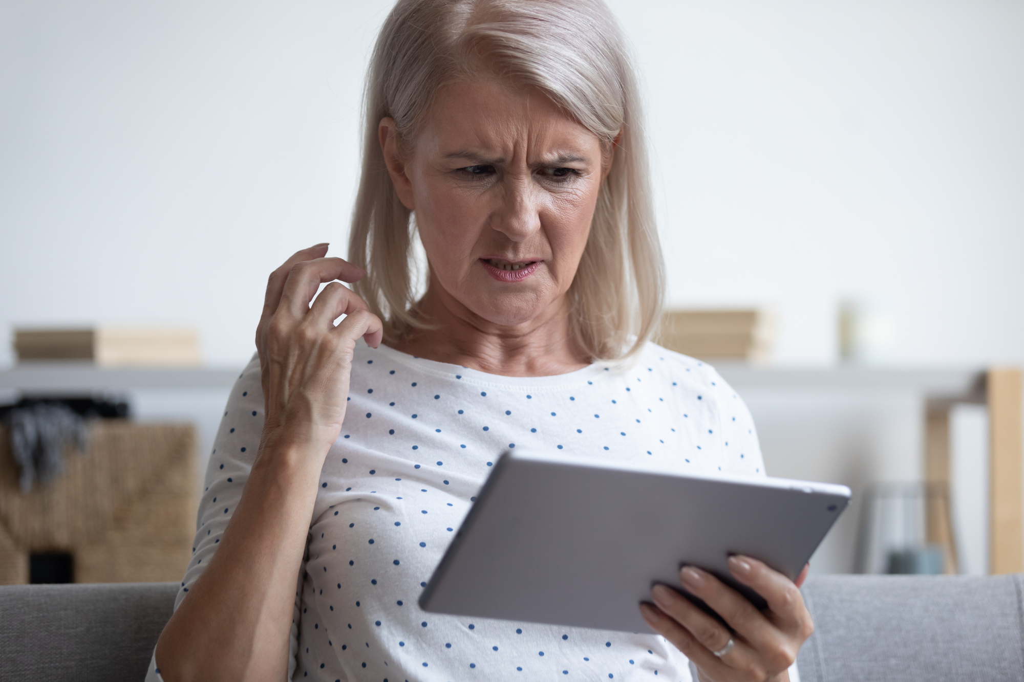 An elderly woman with shoulder-length gray hair, wearing a white shirt with blue polka dots, is seated and holding a tablet. She looks puzzled and is scratching her head as she examines the screen. The background shows a softly blurred indoor setting.