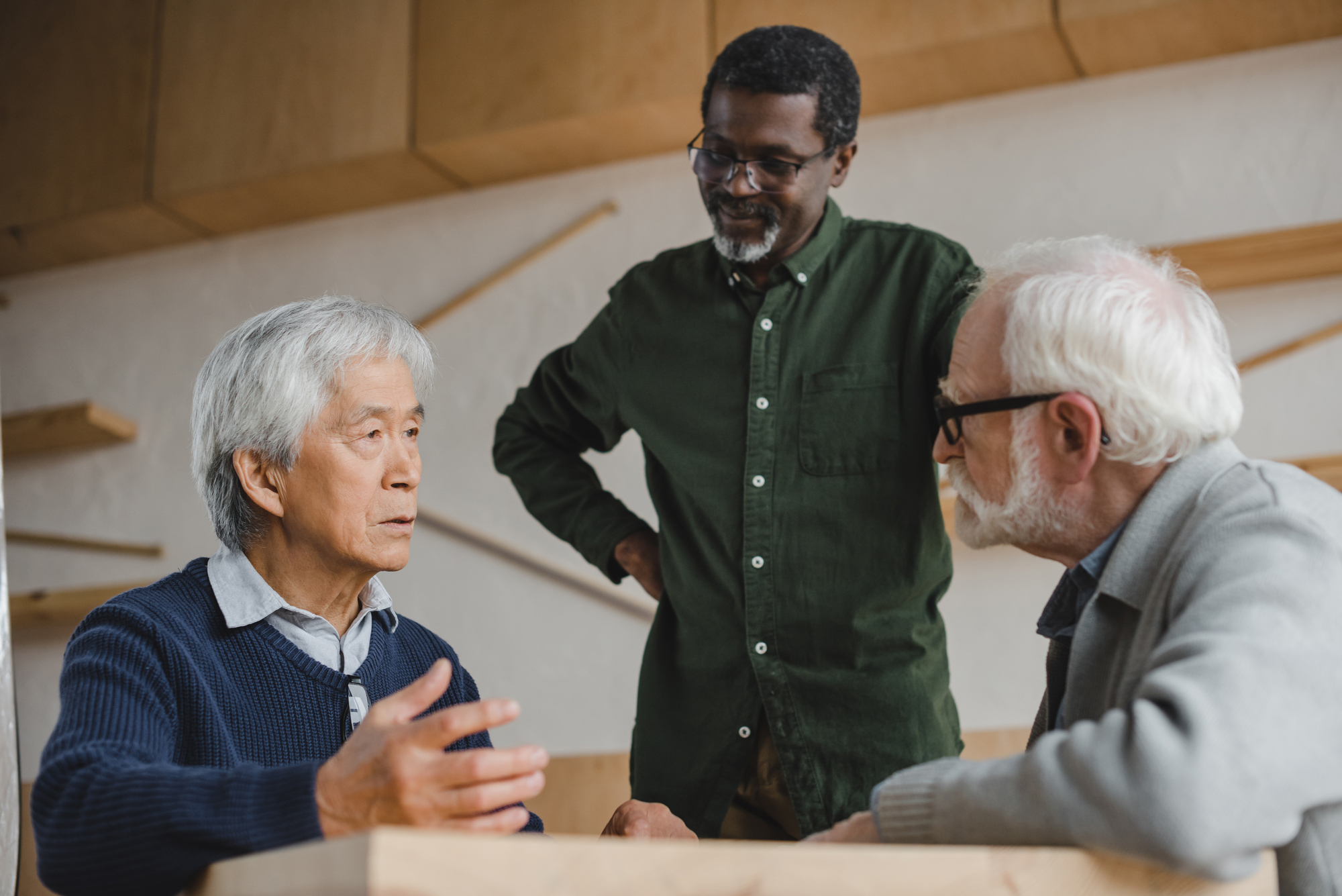 Three elderly men engage in a serious conversation in a modern room with wooden accents. One man with white hair gestures while talking, another with glasses listens intently, and a third man stands between them observing the exchange.