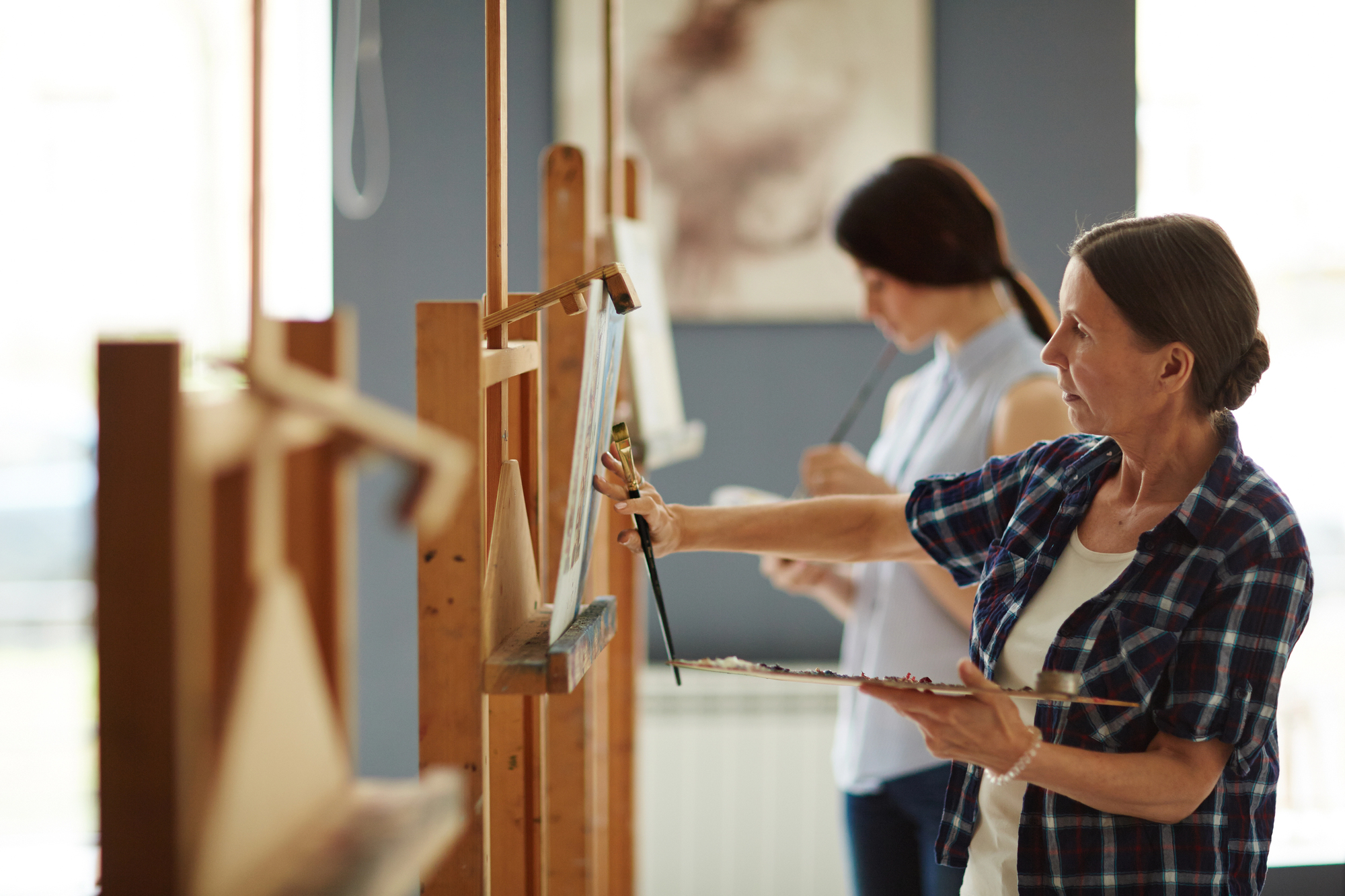 Two women painting at easels in an art studio. The woman in the foreground, wearing a checkered shirt, focuses intently on her canvas, brush in hand, and palette in the other. The woman in the background, in a sleeveless shirt, works on her painting.