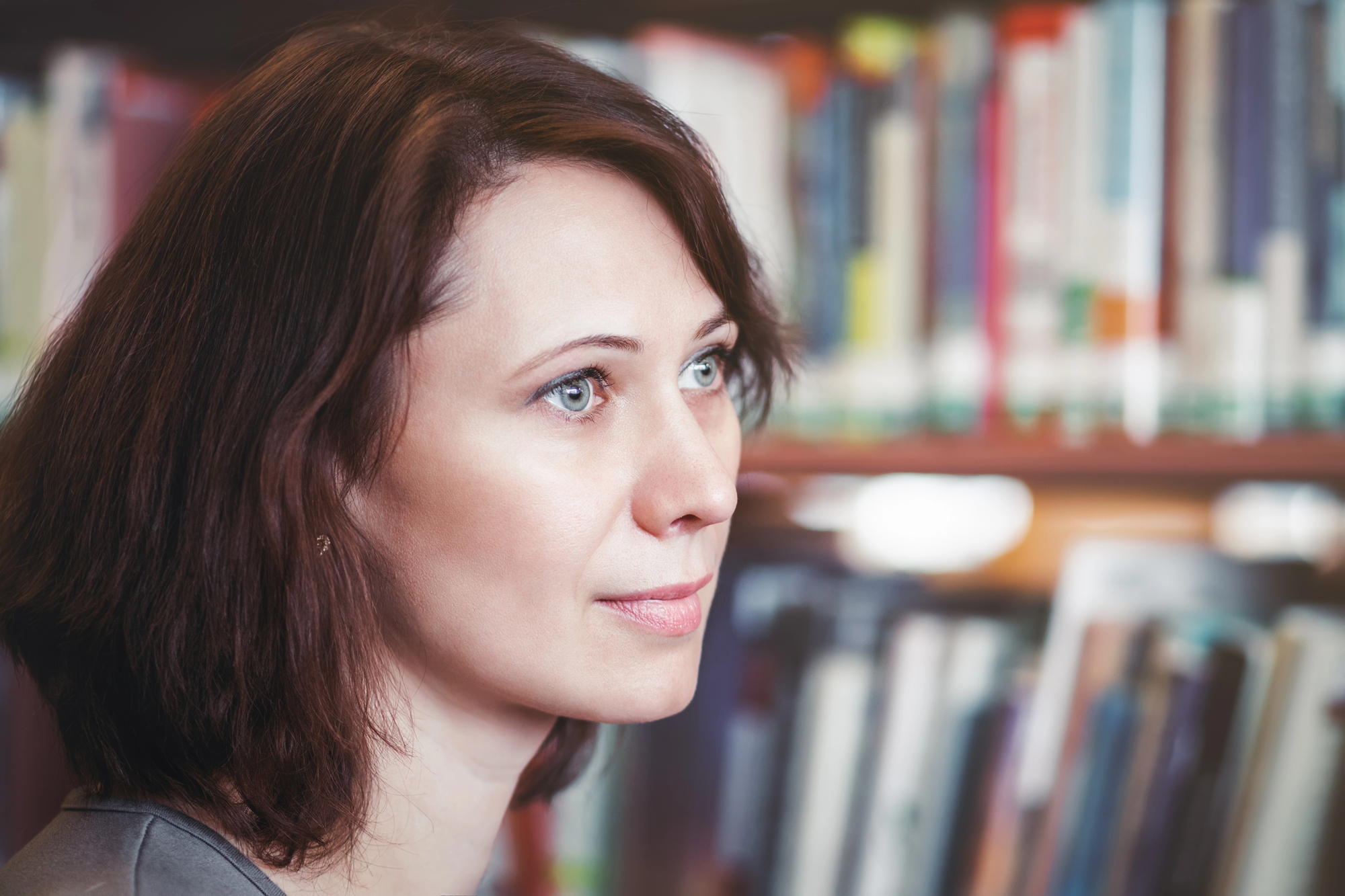 A woman with shoulder-length brown hair and light-colored eyes is gazing thoughtfully to her right. She is in a library, with shelves of colorful books out of focus in the background.