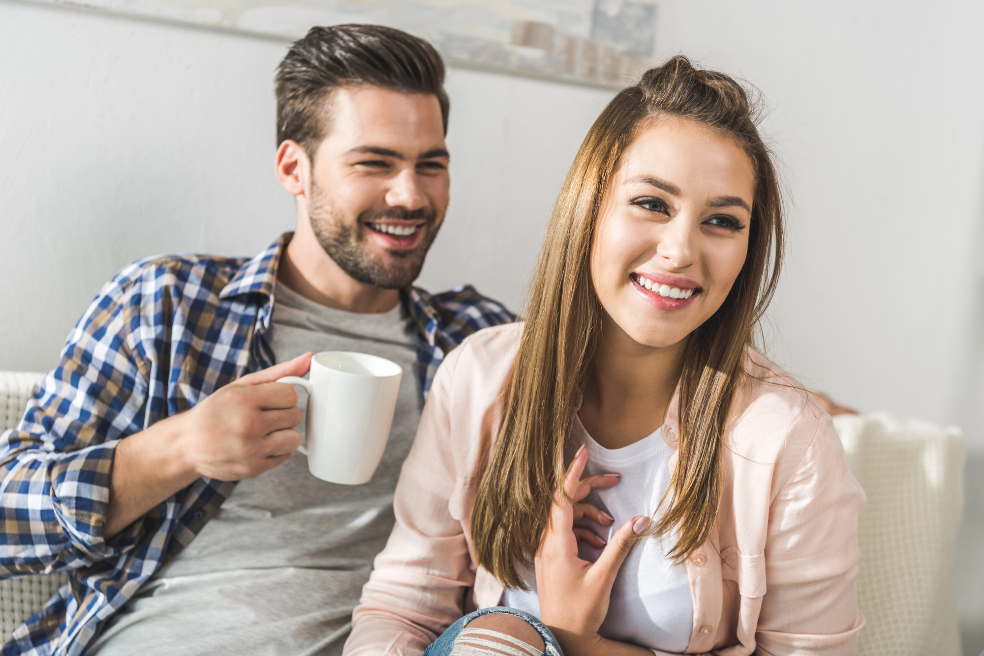 A man and a woman are sitting together on a couch, smiling and looking content. The man, wearing a checkered shirt, is holding a white mug, while the woman, wearing a light pink shirt, is resting her hand near her neck. Both appear relaxed in a bright, cozy setting.