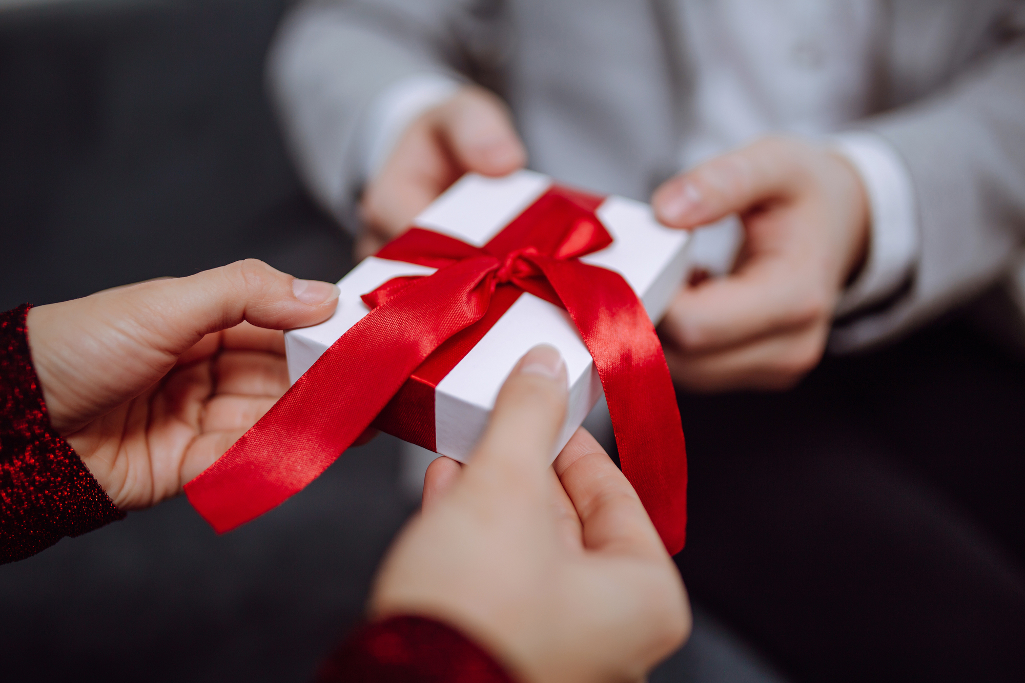 Close-up of two pairs of hands exchanging a gift wrapped in white paper with a red ribbon. One person is wearing a sparkly red sleeve, and the other person is in a light-colored suit. The focus is on the gift being passed between them.