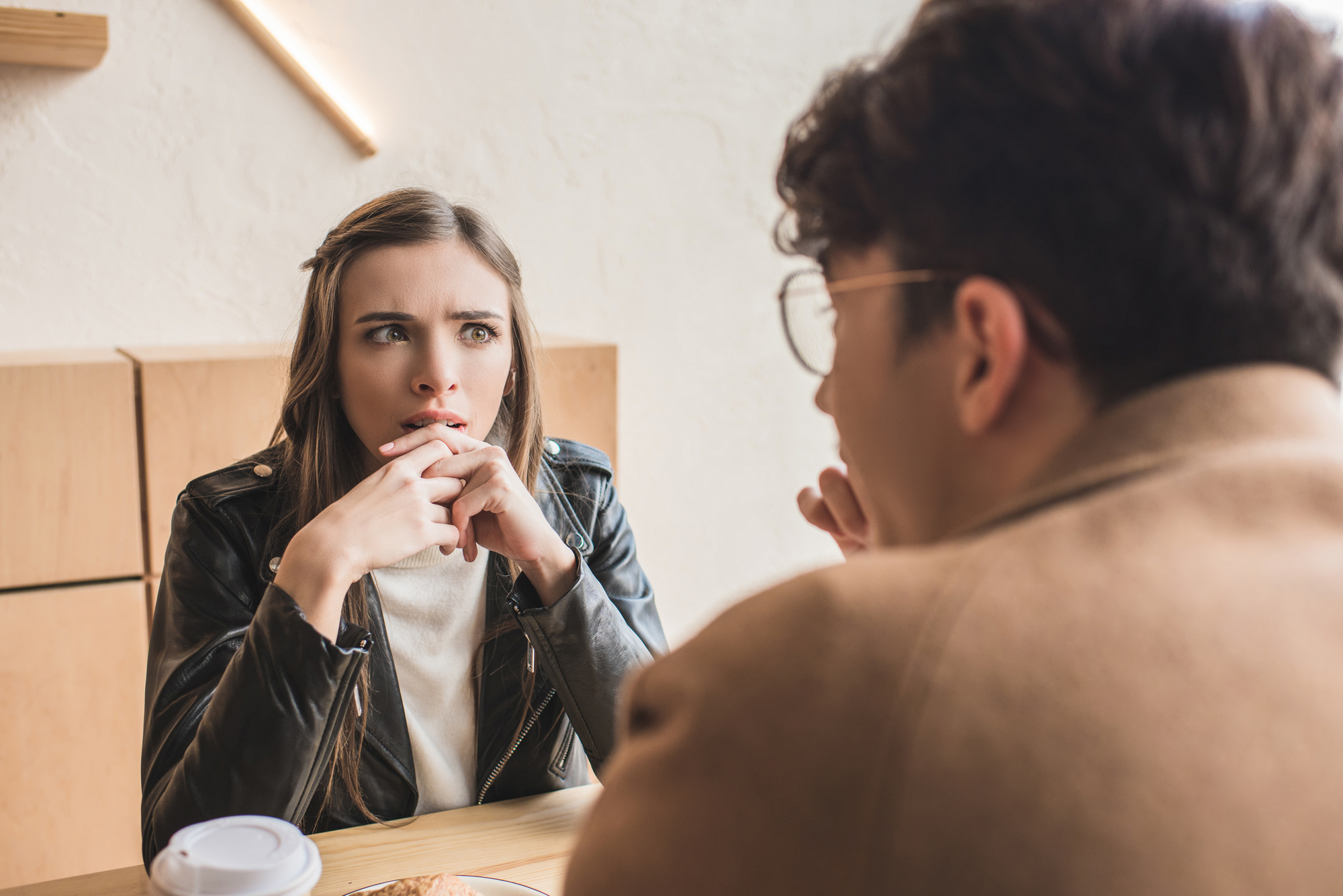 A woman with long brown hair in a leather jacket sits at a table, looking concerned with her hands near her mouth. She faces a person with curly hair and glasses, who is turned away from the camera. They are indoors, with a cup of coffee on the table.