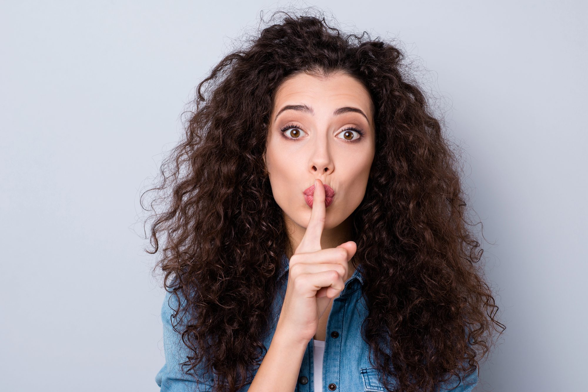 A woman with long, curly hair is standing in front of a plain background, looking directly at the camera with wide eyes. She is wearing a denim shirt and holding her index finger up to her lips in a shushing gesture.