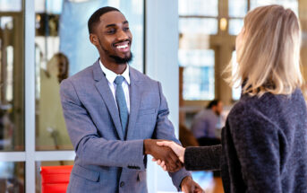 A man in a grey suit and tie, smiling, shakes hands with a woman in a dark blazer. They are standing in a modern office setting with large glass windows in the background. The man holds a piece of paper in his other hand.