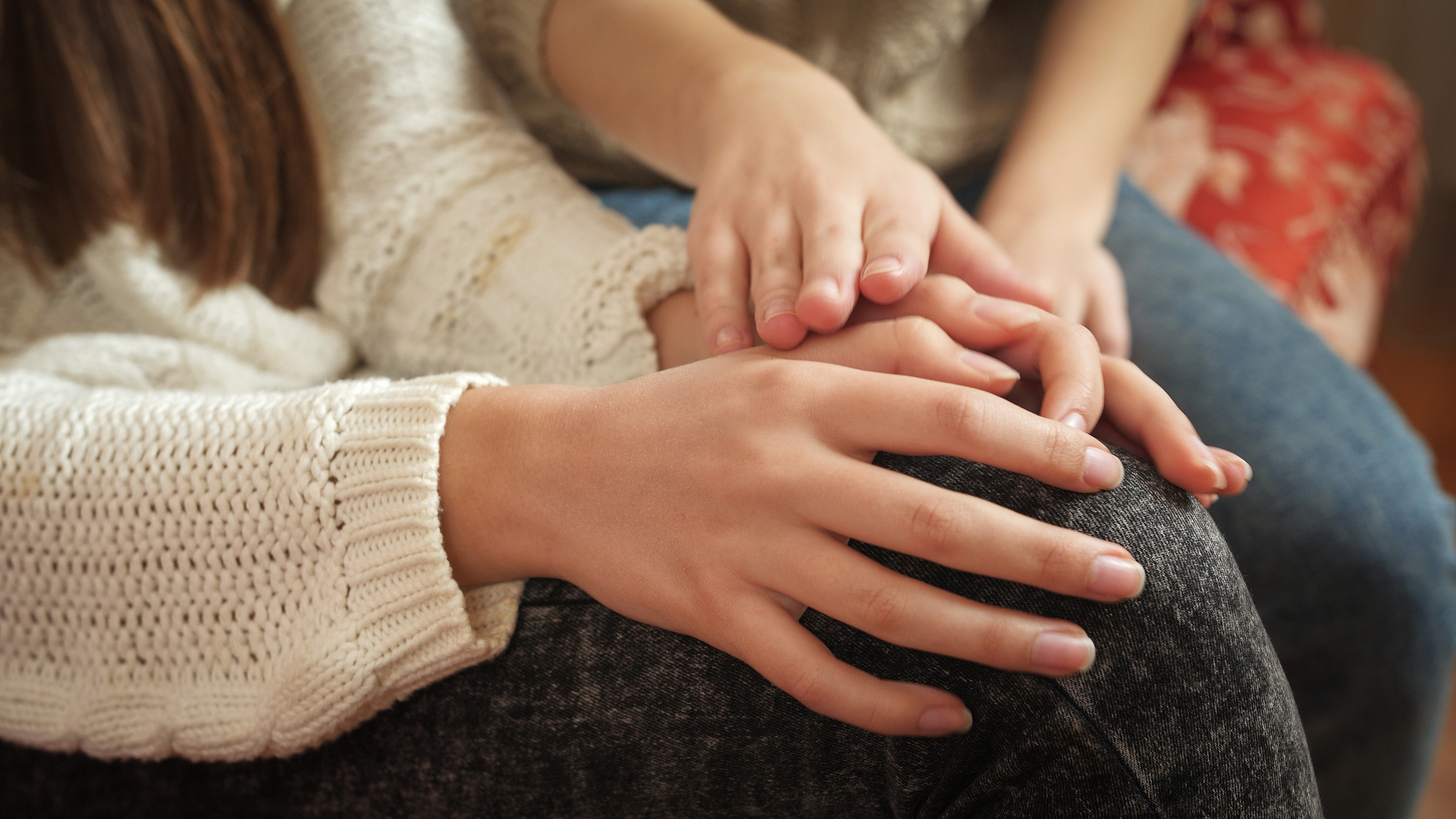 A close-up of two people sitting closely, each placing their hands over the other's. The person on the right gently touches the hand of the other, who is wearing a white knitted sweater and black pants. The image conveys a sense of comfort and support.