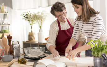 An elderly woman wearing glasses and a red apron and a young woman in a striped shirt are making dough together in a kitchen. Various cooking ingredients and utensils, including a recipe book, are on the countertop. A potted plant and kitchen tools are in the background.