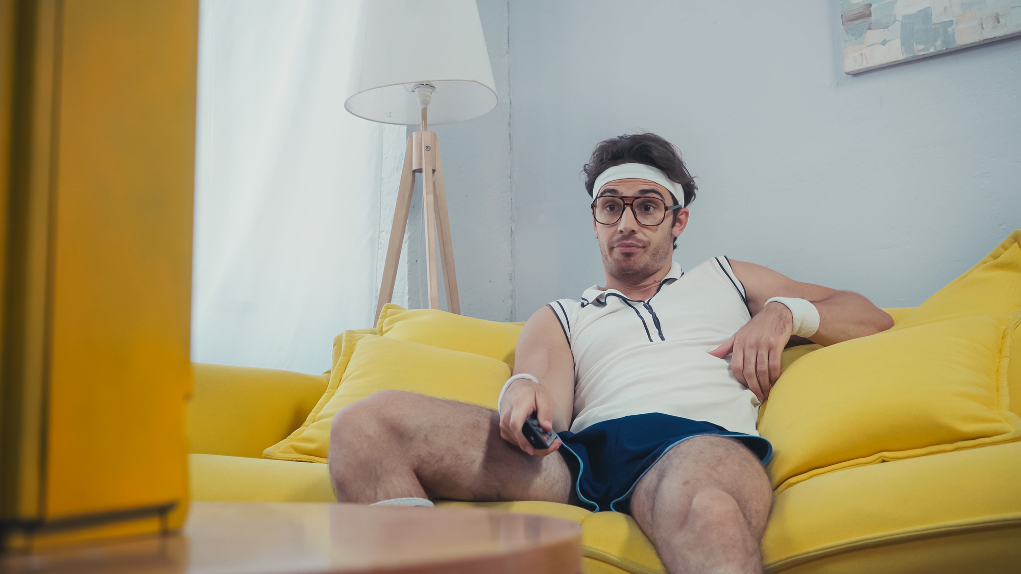 A man dressed in retro athletic attire, including a headband, wristbands, glasses, and shorts, is lounging on a yellow sofa with a remote control in hand, watching TV. He has a relaxed and somewhat amused expression on his face.