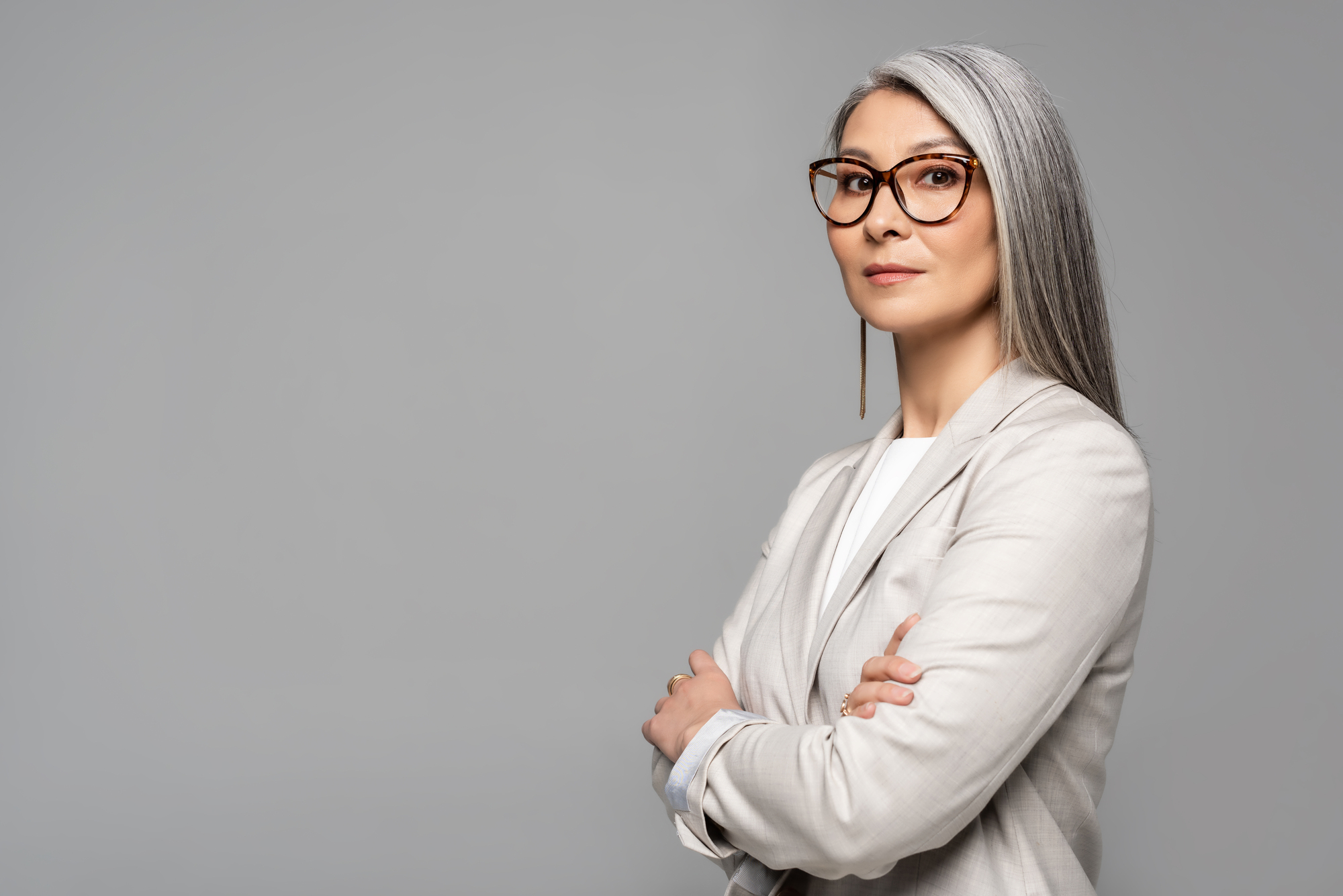 A confident woman with long gray hair wears glasses and a light-colored blazer. She stands with her arms crossed, looking directly at the camera against a plain gray background.