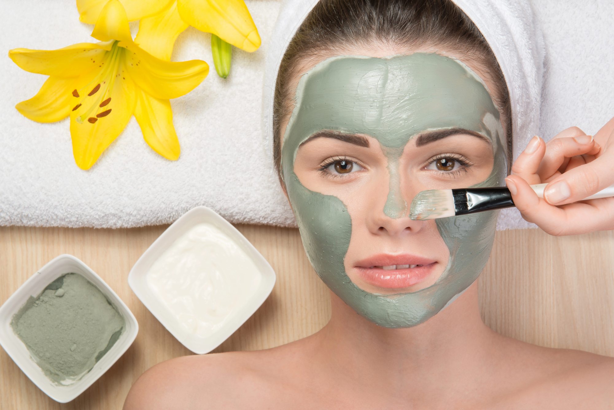 A woman is receiving a facial treatment, lying on a towel with two yellow lilies nearby. A hand is applying a green face mask to her face with a brush. Two small bowls containing different colored face masks are placed next to her.