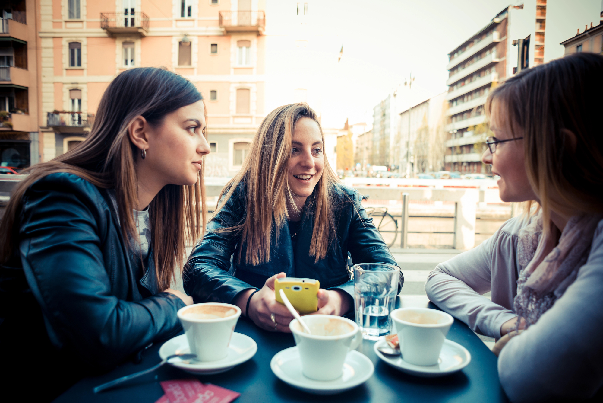 Three young women sit at an outdoor café table, engaged in a lively conversation. Each has a cup of coffee in front of them. The woman in the middle is holding a yellow smartphone and smiling. Urban buildings are visible in the background.