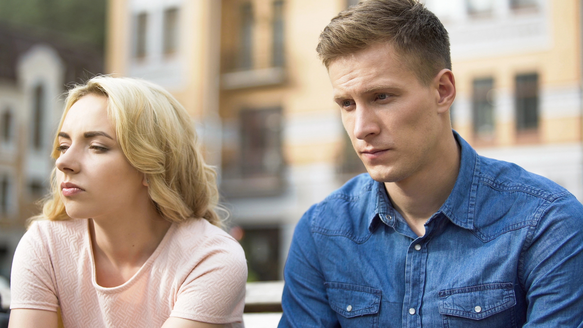 A man and woman sit side by side outdoors, both appearing troubled and deep in thought. The woman, in a pink top, looks away to the left, while the man, in a blue button-down shirt, gazes downward. The background shows a blurred urban setting with buildings.