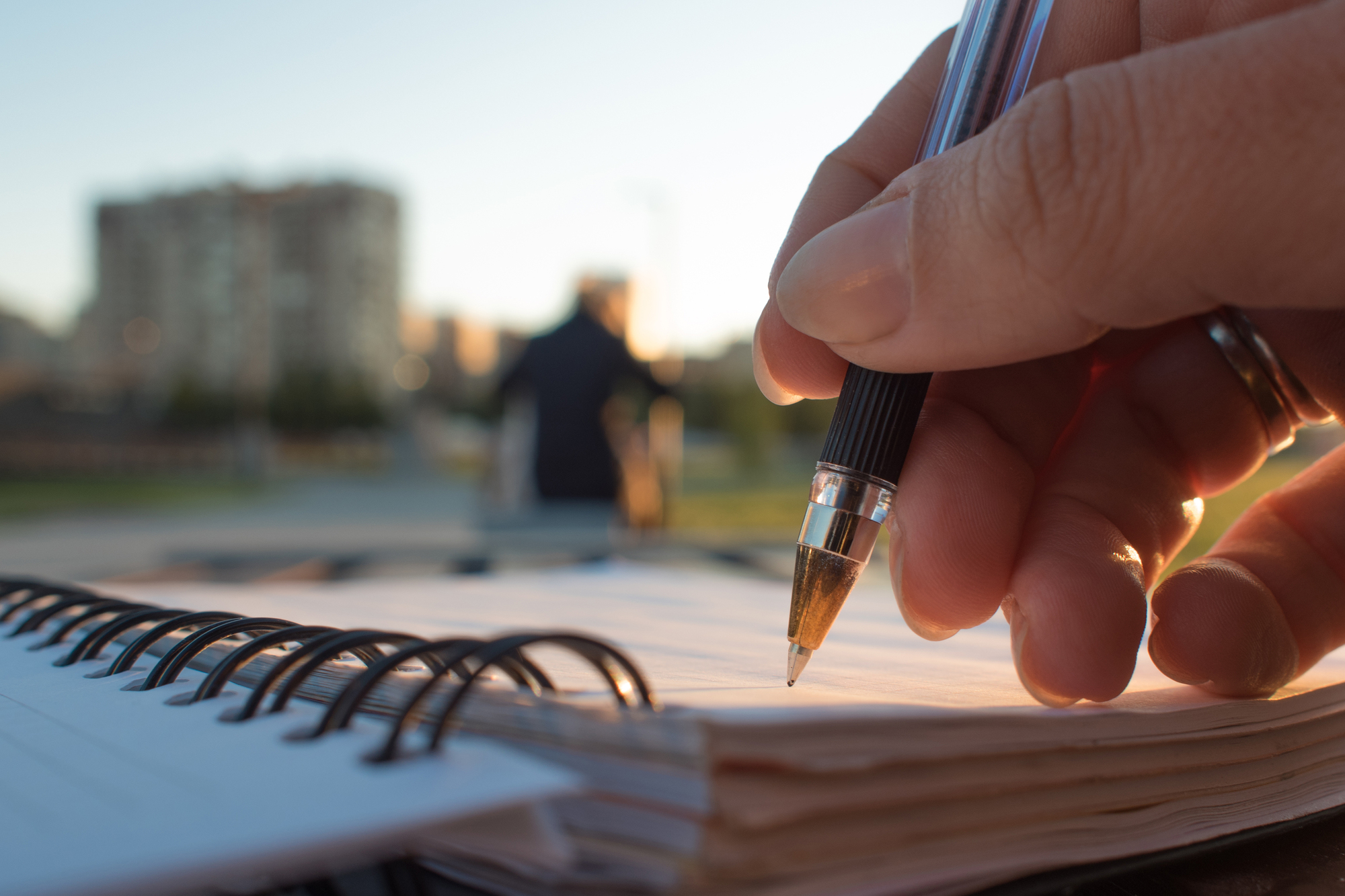 A close-up of a hand holding a pen and writing on a spiral-bound notebook. The scene is set outdoors with soft sunlight, and a blurred background featuring a person sitting on a bench and buildings in the distance.