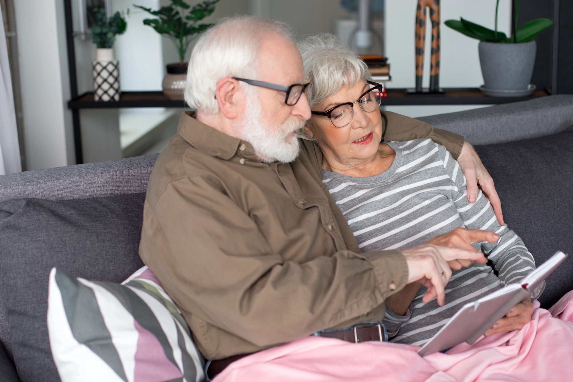 An elderly couple sits on a couch, cozily wrapped in a pink blanket. The man, wearing glasses and a brown shirt, points at a tablet, while the woman, also wearing glasses and a striped shirt, leans in and looks at the screen intently. Houseplants are visible in the background.