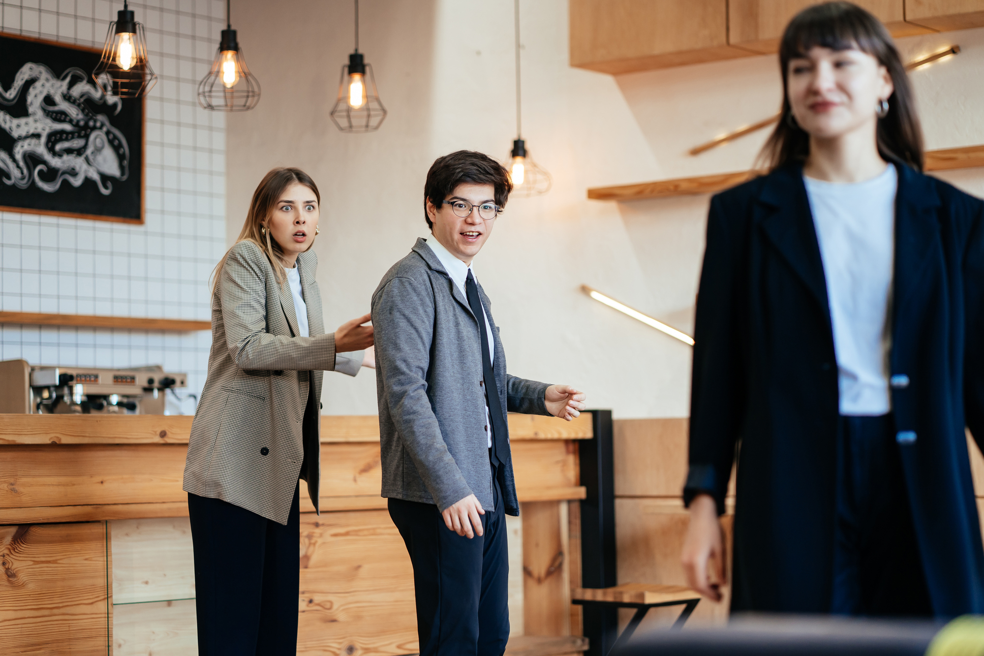 Three people in a modern café. A woman with blonde hair stands behind and gestures towards a person in a gray blazer and glasses who looks startled. Another woman in a dark outfit is walking away in the foreground, slightly blurred. Hanging lights and wooden decor are visible.