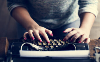 A person with red-painted nails types on a vintage typewriter. The paper in the typewriter is blank, and their face is not visible. They wear a gray, long-sleeved shirt. The atmosphere suggests a focus on writing or creativity.