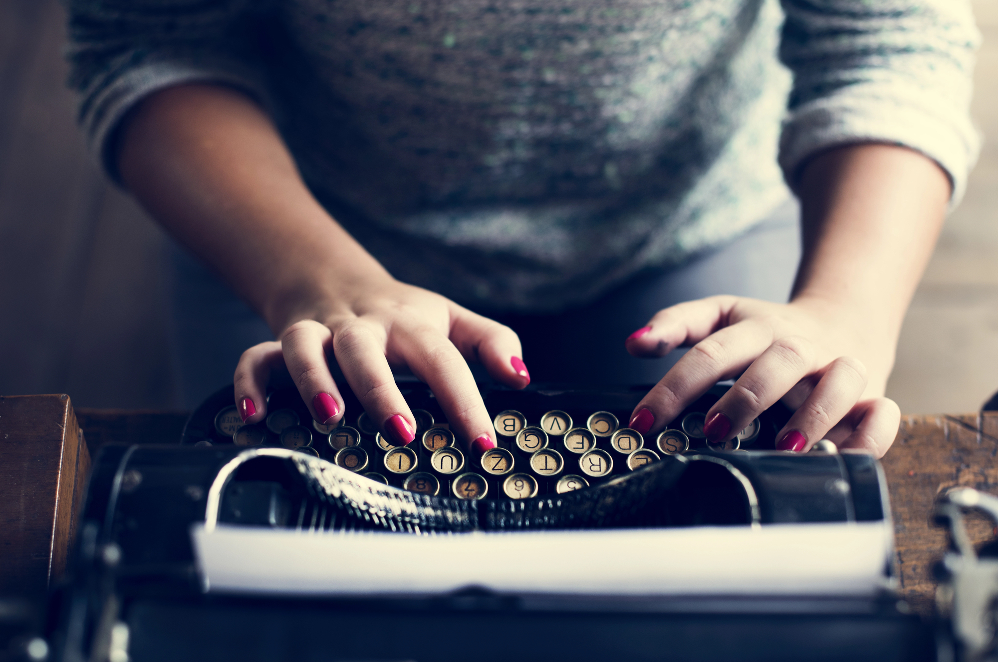 A person with red-painted nails types on a vintage typewriter. The paper in the typewriter is blank, and their face is not visible. They wear a gray, long-sleeved shirt. The atmosphere suggests a focus on writing or creativity.