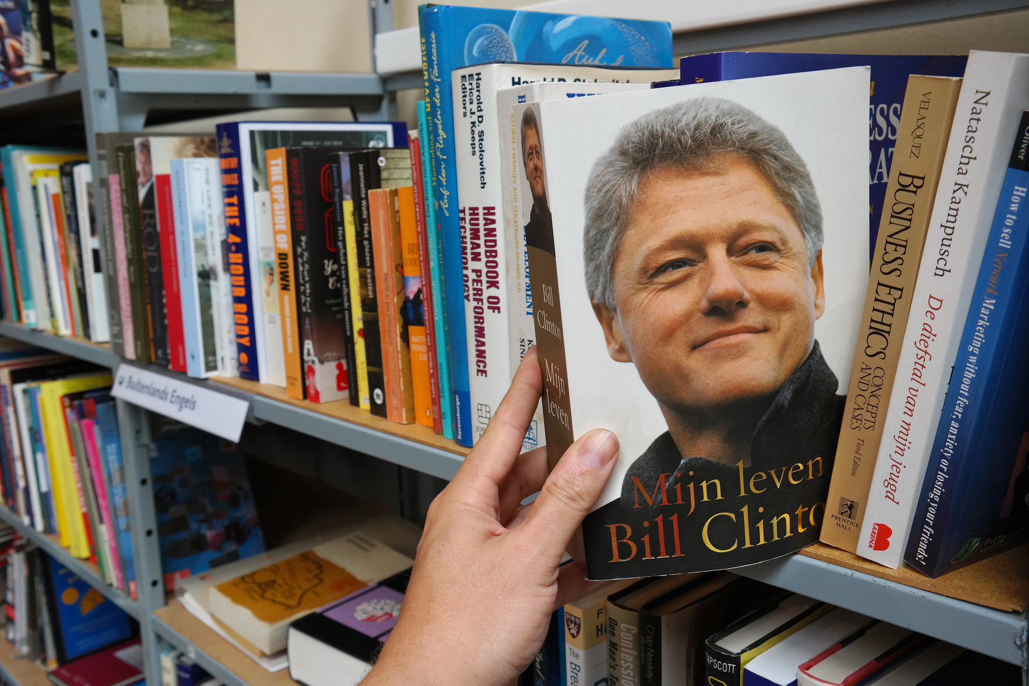 A person is holding a book titled "Mijn leven" by Bill Clinton on a bookshelf. The shelf is filled with various books in different languages and genres. One section on the shelf is labeled "In verschillende Engeln" indicating multiple books in English.