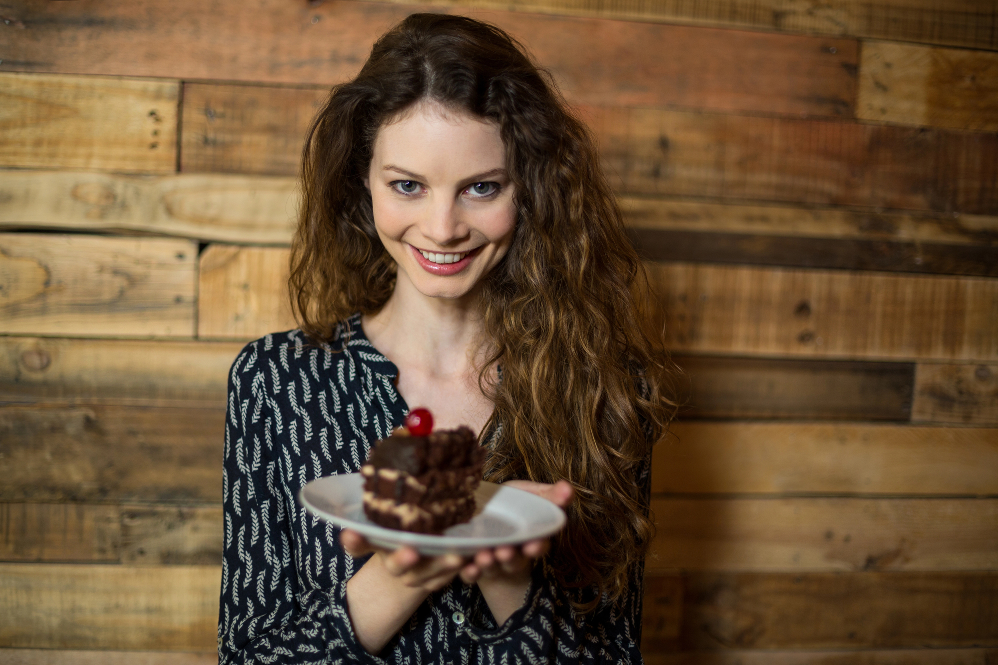 A woman with long curly hair smiles while holding a plate with a piece of chocolate cake topped with a cherry. She is standing against a wooden plank background, wearing a patterned black and white blouse.