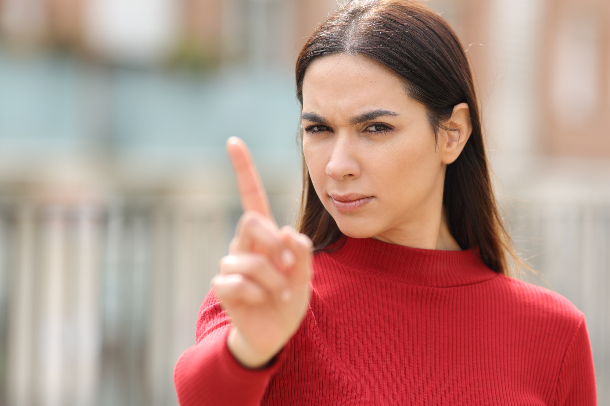 A woman with long dark hair wearing a red sweater is standing outdoors and looking directly at the camera with a serious expression. She is holding up her index finger as if to indicate "no" or "stop." The background is blurred.